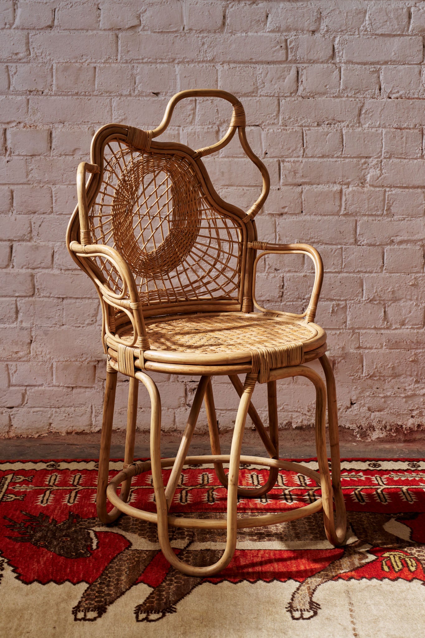 Wicker armchair with a curved back and intricate weaving pattern, featuring natural rattan material and a polished finish, placed on a patterned rug against a brick wall.