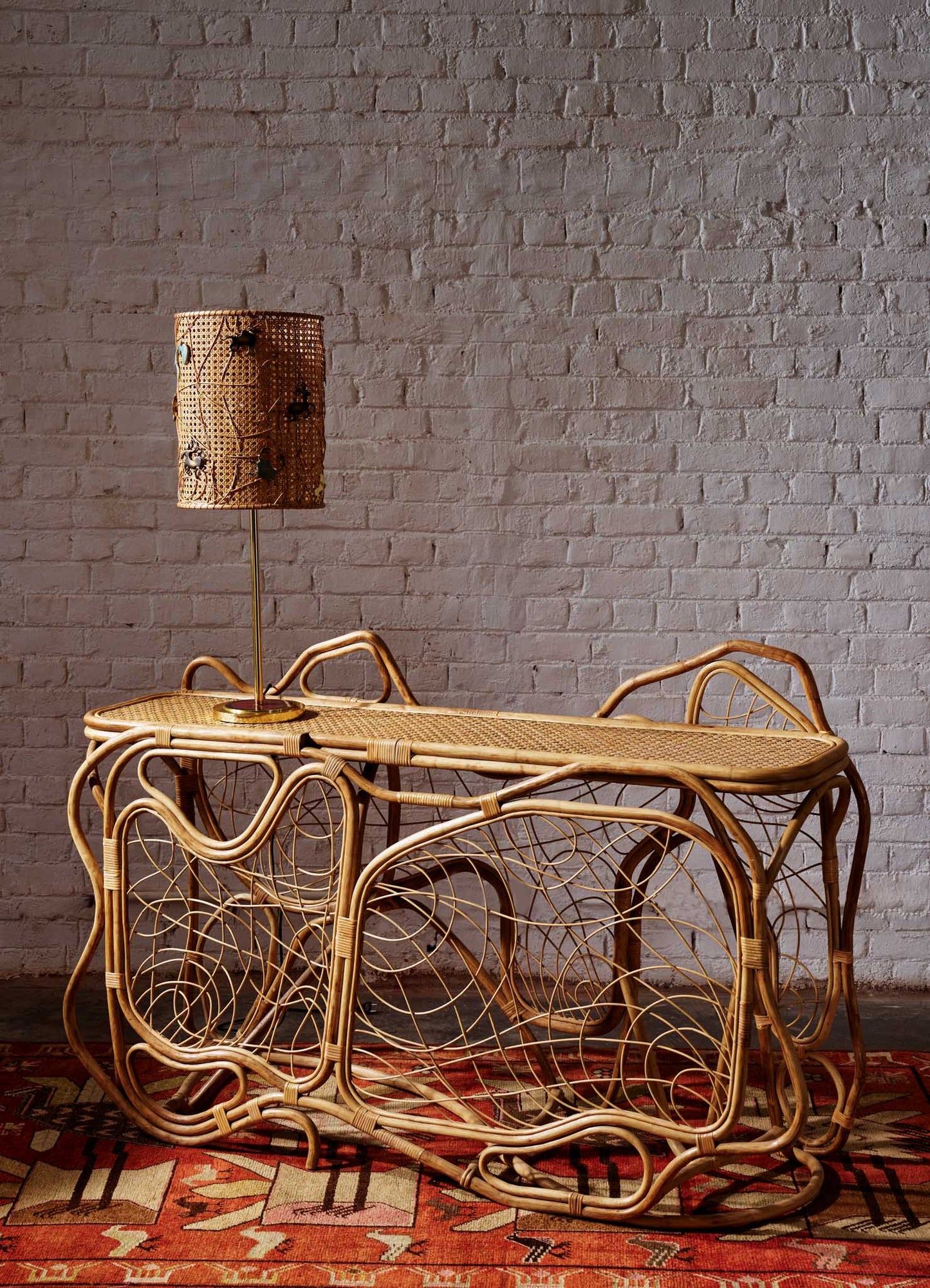 Rattan console table with an intricate, swirling pattern and a natural wood finish, paired with a matching rattan table lamp featuring a woven shade, set against a white brick wall. The scene includes a geometric-patterned area rug with earthy tones.