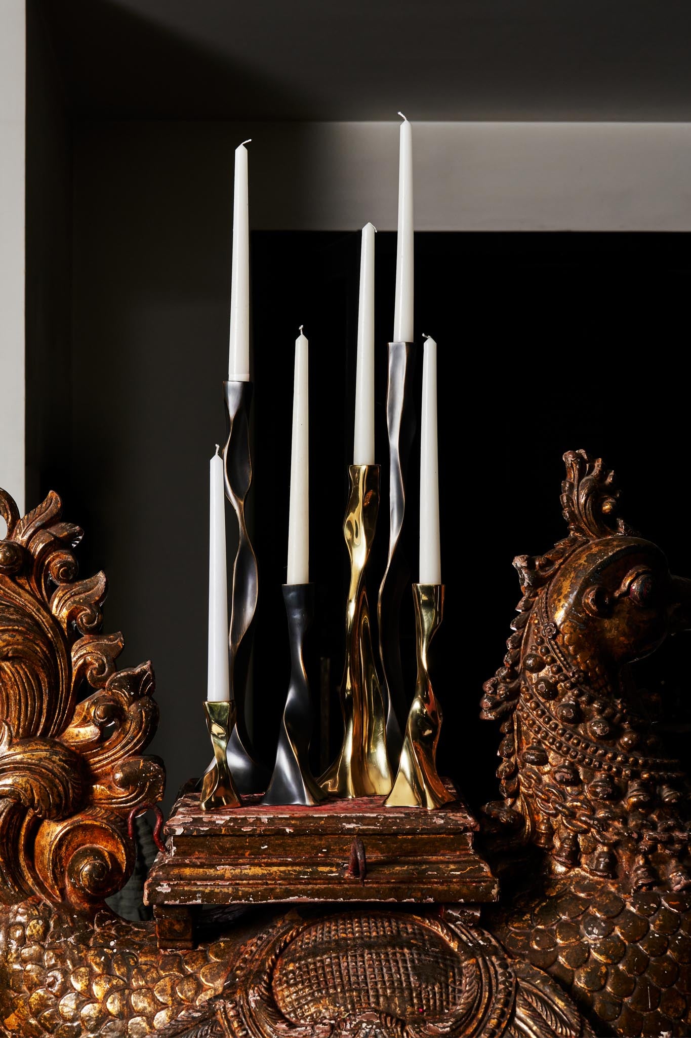 A set of tall, twisted metal candlesticks with a polished finish in gold, silver, and black, holding white taper candles. They are arranged on a distressed wooden surface, flanked by ornate wooden carvings with intricate patterns and a rich, dark finish.