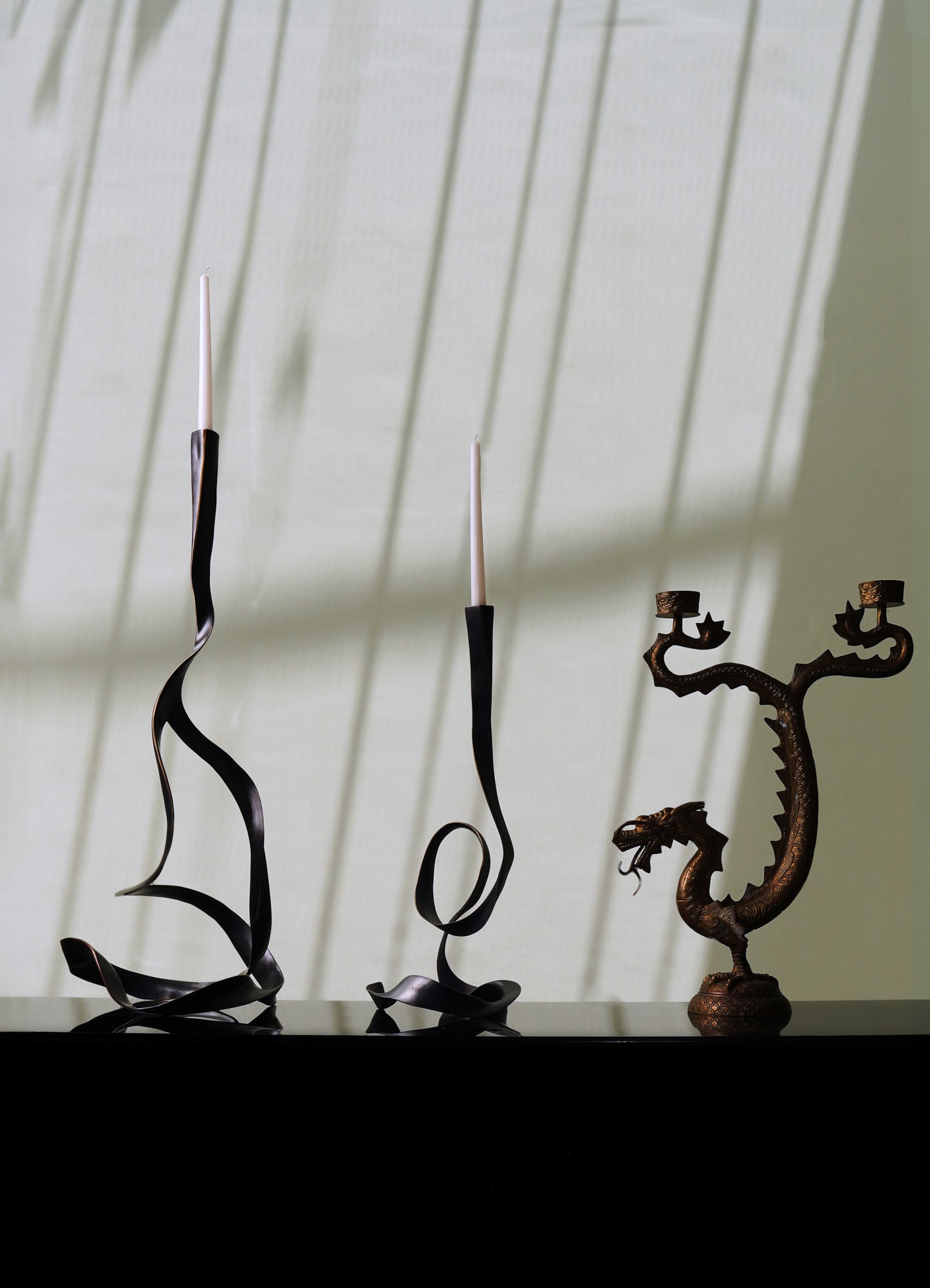 Black metal candleholders with a twisted, sculptural design holding white taper candles, along with a decorative bronze dragon motif sculpture with intricate detailing, set against a light green wall with geometric shadows.