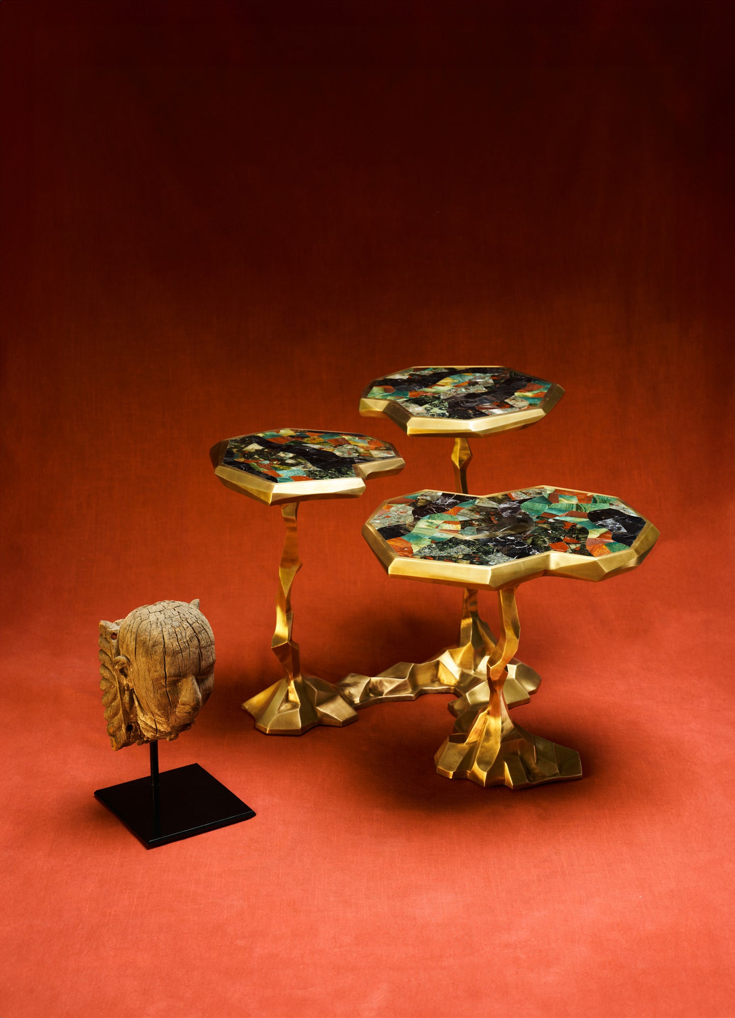A set of three decorative side tables with irregular, sculptural gold-finished metal bases and octagonal tops featuring multicolored mosaic patterns. The tables are displayed on a red background along with a carved wooden head sculpture mounted on a black stand.