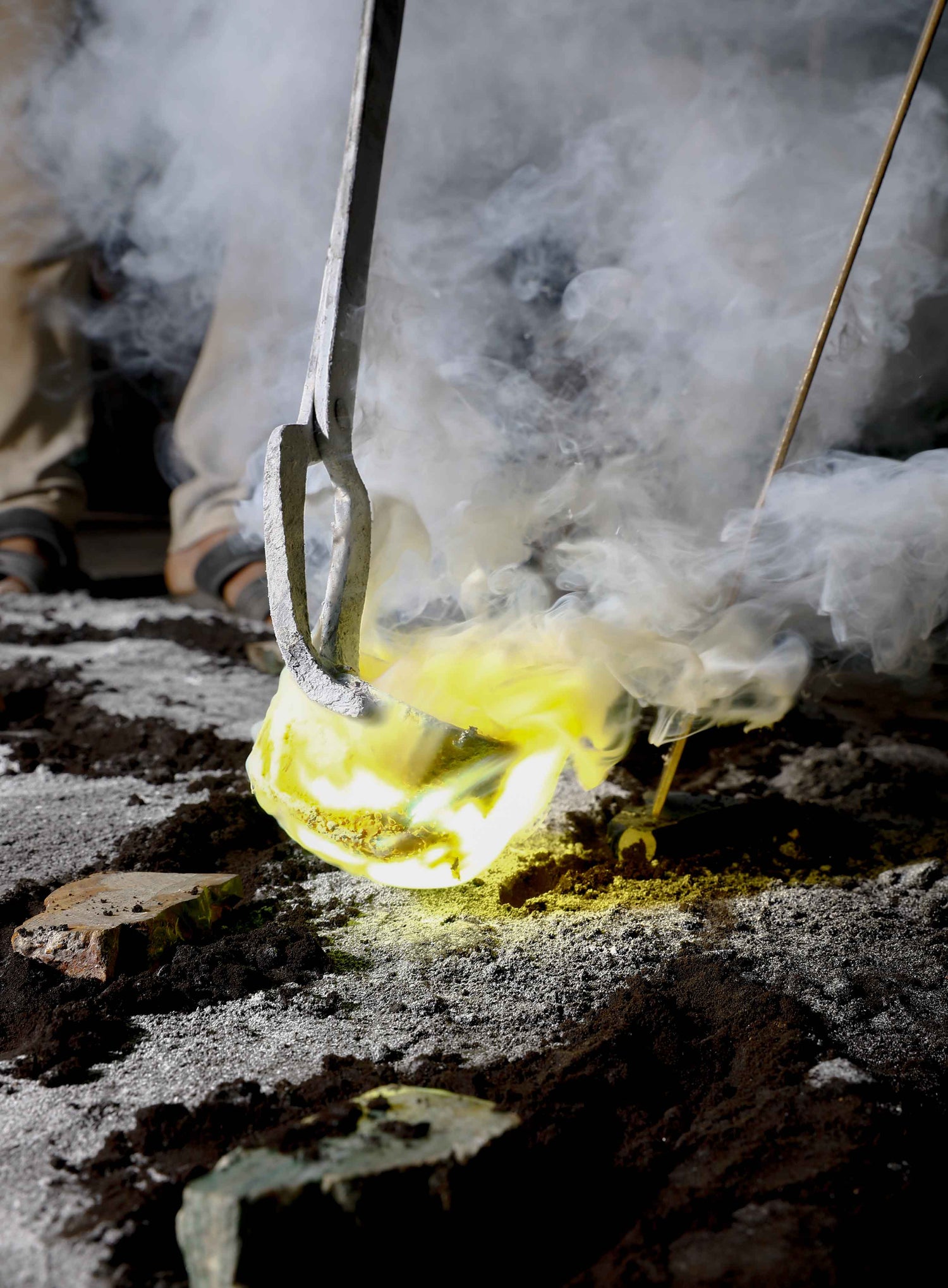A glass blower working with molten glass, creating a glowing yellow-orange glass object amidst billowing smoke and steam, with tools and hands visible in the process.