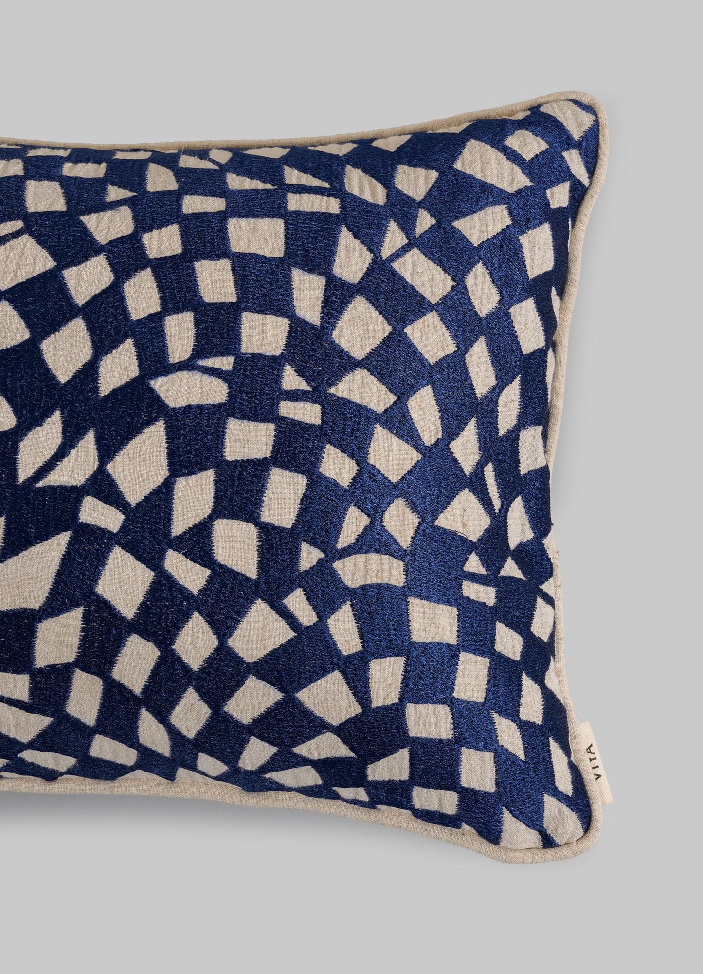 The Viya Dreamscape Blue Embroidered Checks Cushion Cover 12"x18" features blue and beige geometric patterns on a light gray background, accented with a small fabric tag on the side.