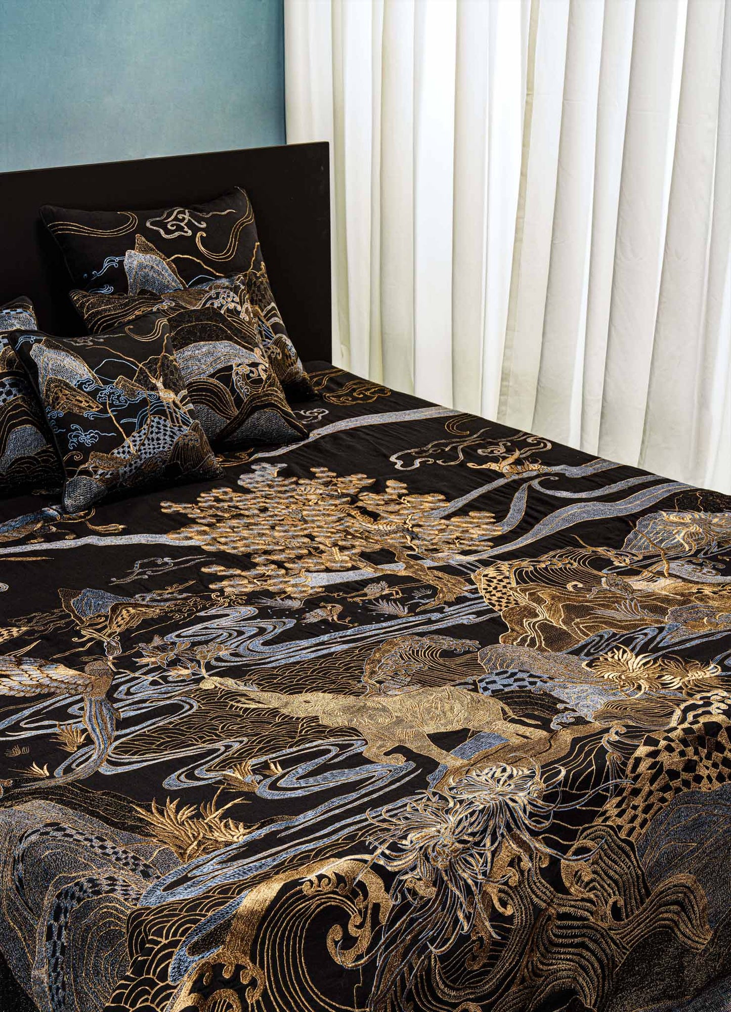 A neatly made bed with the Viya Dreamscape Black & Gold Embroidered Bedcover and matching pillows featuring nature-inspired motifs, set beside softly lit white curtains.