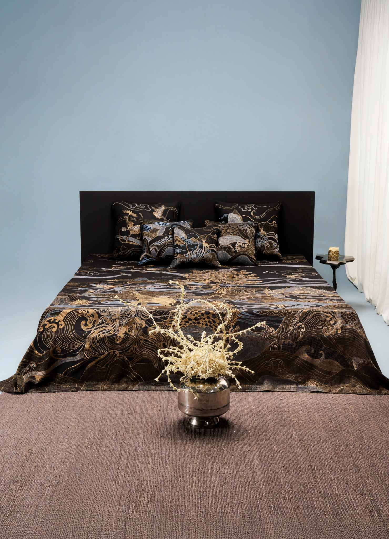 A modern bedroom showcases Viya's Dreamscape Black & Gold Embroidered Bedcover on a black bed frame, paired with patterned pillows, a small side table with a cup, sheer white curtains, and dried flowers on a brown rug.