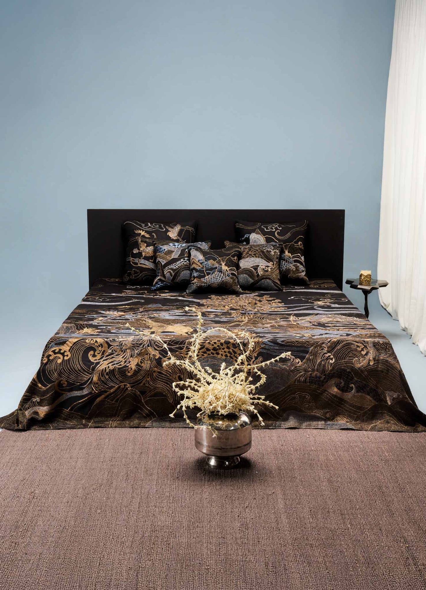A bed with black bedding featuring gold embroidered floral and wave motifs, accompanied by matching decorative pillows. The room has a black headboard, a dark side table with a small decorative object, and a light-colored curtain on the right side. A beige carpet covers the floor.