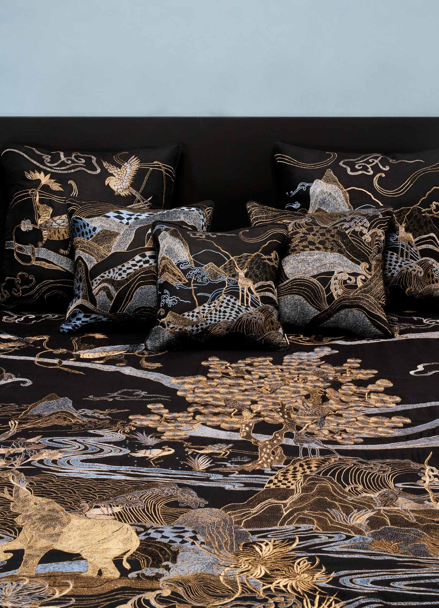 The Viya Dreamscape Black & Gold Embroidered Bedcover, featuring symbolic animal and tree motifs in gold and silver, dresses the bed with matching pillows, set against a plain light wall.