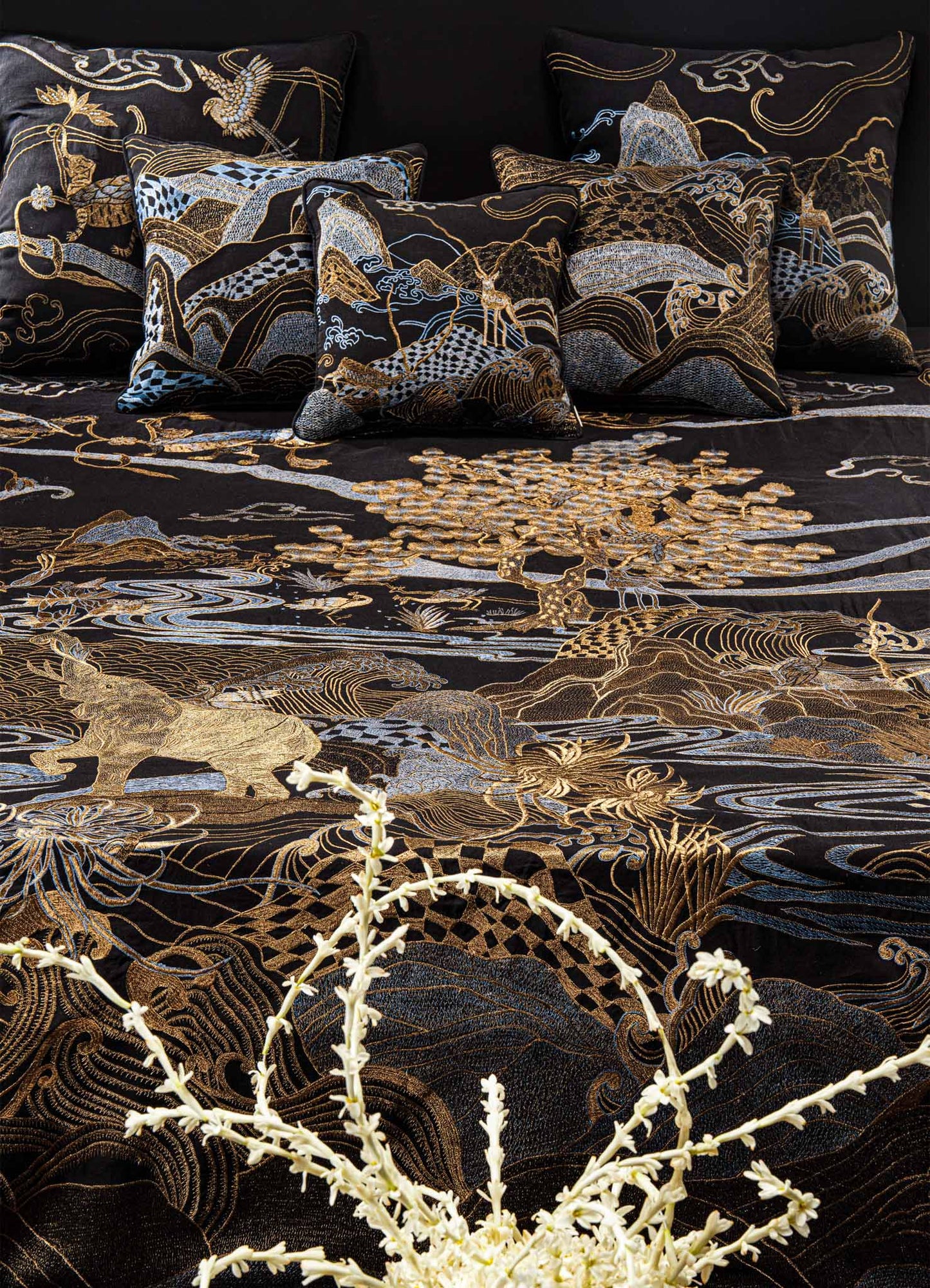 The Viya Dreamscape Black & Gold Embroidered Bedcover features intricate blue and gold embroidery with animal and nature motifs, paired with matching pillows and a white floral arrangement in the foreground.