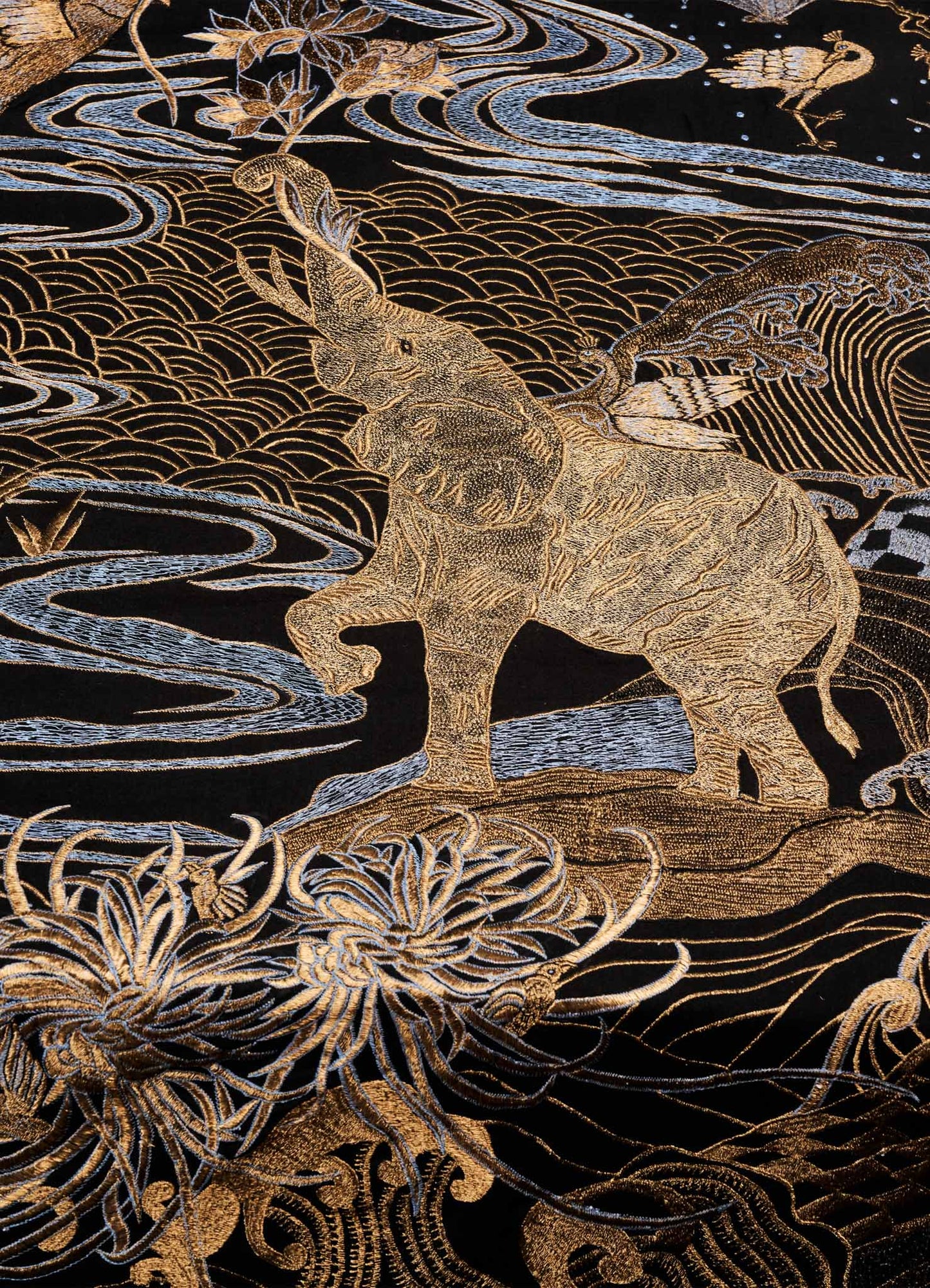 The Viya Dreamscape Black & Gold Embroidered Bedcover features intricate gold embroidery on dark fabric, depicting a raised-trunk elephant amid swirling water, flowers, and birds for a rich, ornate look.