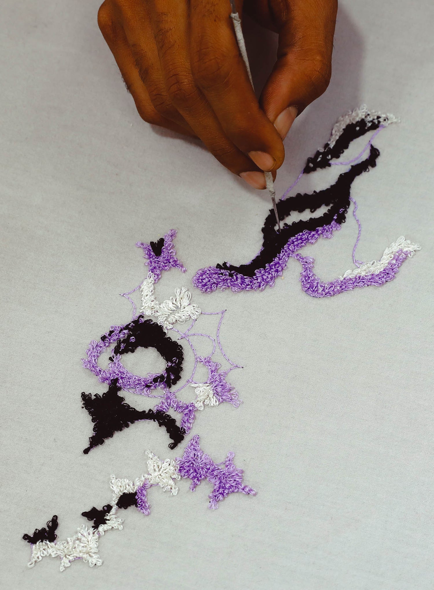 Hand embroidery featuring Halloween-themed motifs, including bats and spiders, in black, white, and purple threads on a fabric surface. The embroidery has intricate, textured stitching creating a decorative and festive pattern.