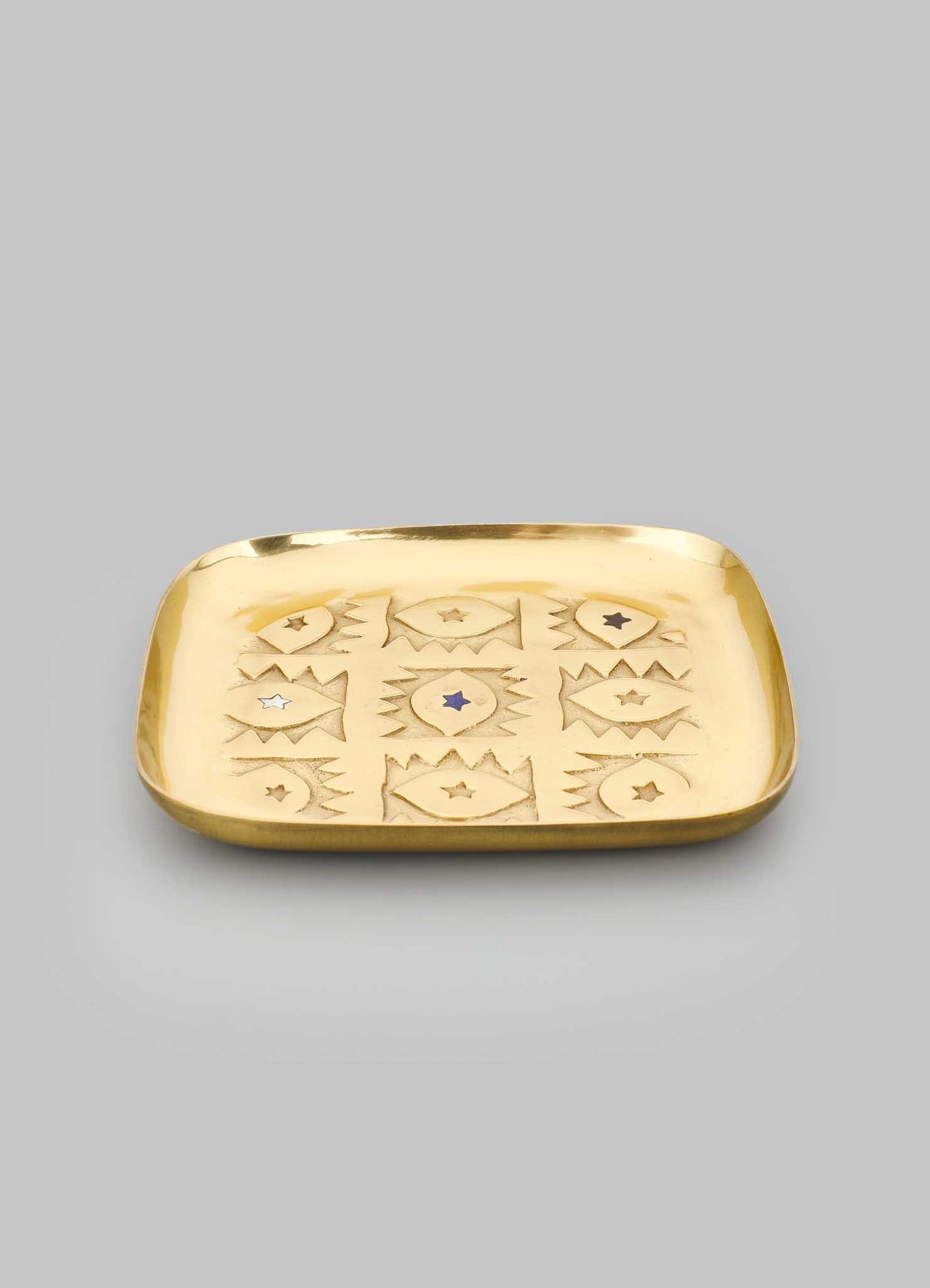 The VIYA Evil Eye Trinket Tray Large is a square, gold-colored metal tray with raised edges and geometric star patterns inspired by Nazar amulets, engraved on its surface and displayed against a plain gray background.