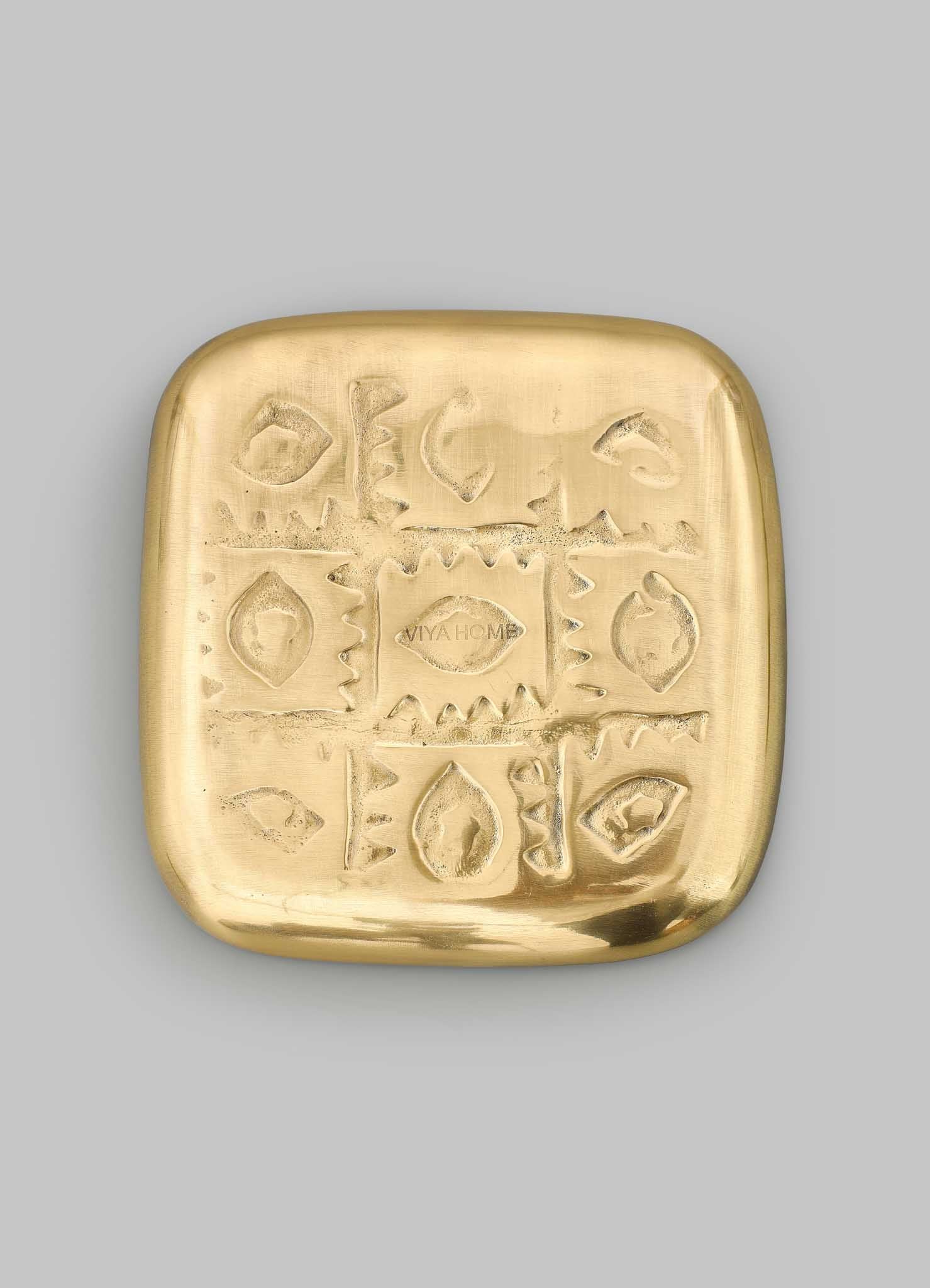 A large, square gold-toned trinket tray by VIYA with engraved geometric and abstract patterns inspired by a Nazar amulet. Photographed on a plain gray background, it features "VIYA" at the center.