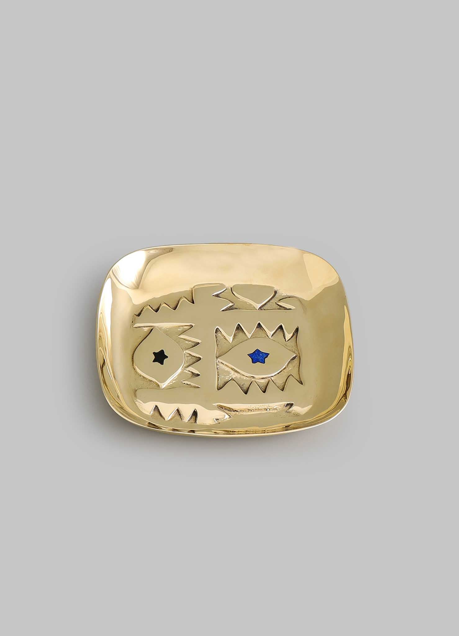 The VIYA Evil Eye Trinket Tray Small is a gold-colored, rectangular dish featuring abstract jagged designs and two star-shaped blue and black inlays, evoking a nazar amulet for subtle evil eye protection. Set against a plain gray background.