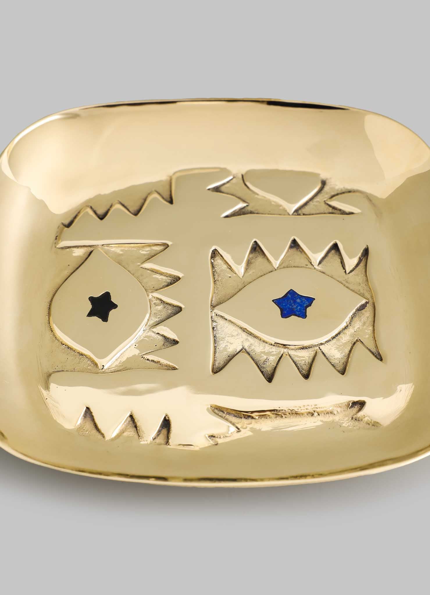 The VIYA Evil Eye Trinket Tray Small is a square, gold-toned dish with abstract engraved eye designs—one with a black star and one with a blue star—serving as a modern home talisman for protection, shown on a gray background.