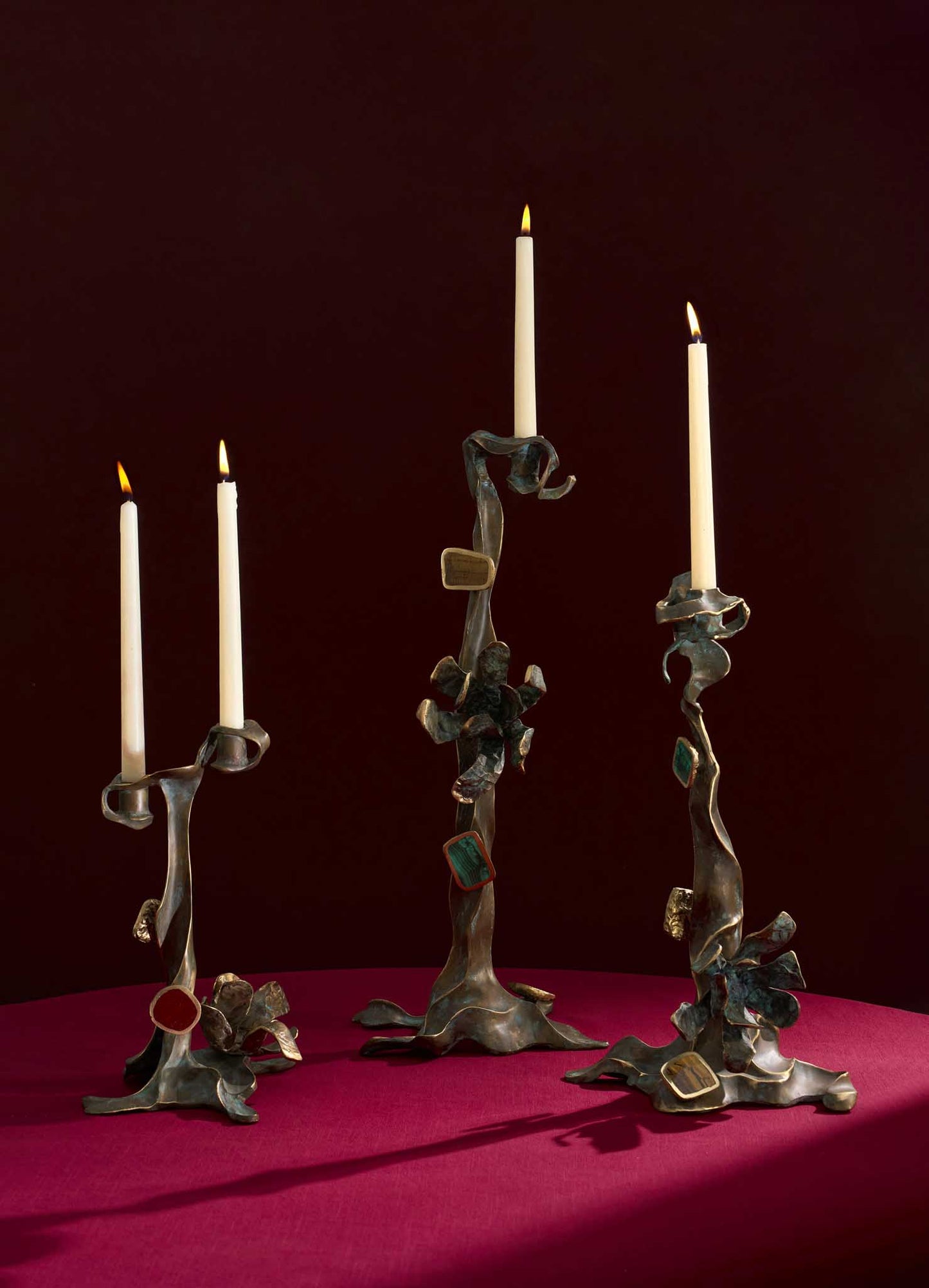The Glory Lily Candle Holder Large by VIYA features sculptural cast brass with twisting, organic forms and colorful accents on magenta, inspired by the medicinal flower. Shown with lit white candles against a dark burgundy background.