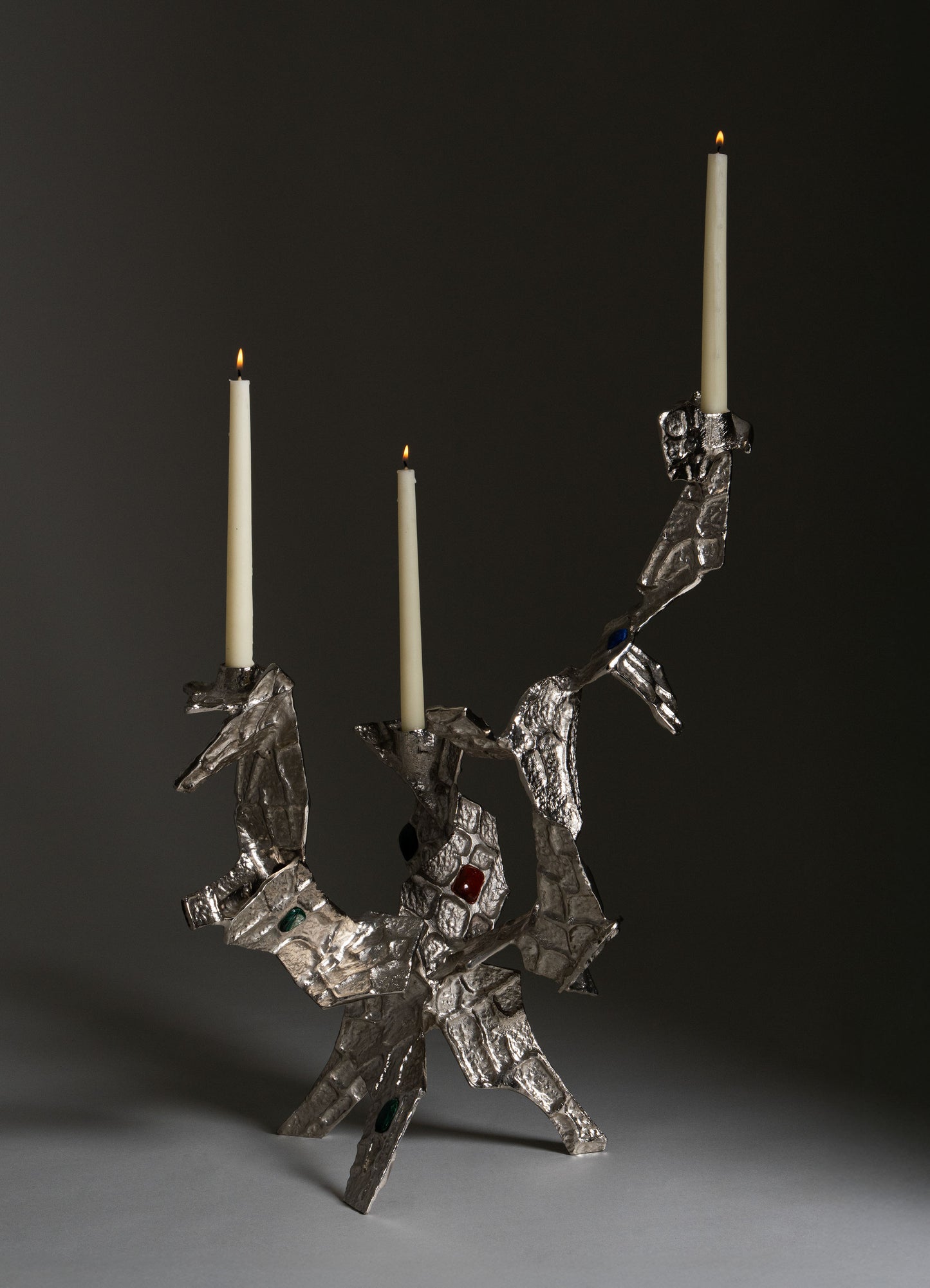 A metallic candlestick holder with an abstract, sculptural design featuring organic, irregular shapes and embedded colored stones, holding three tall white taper candles with lit flames.