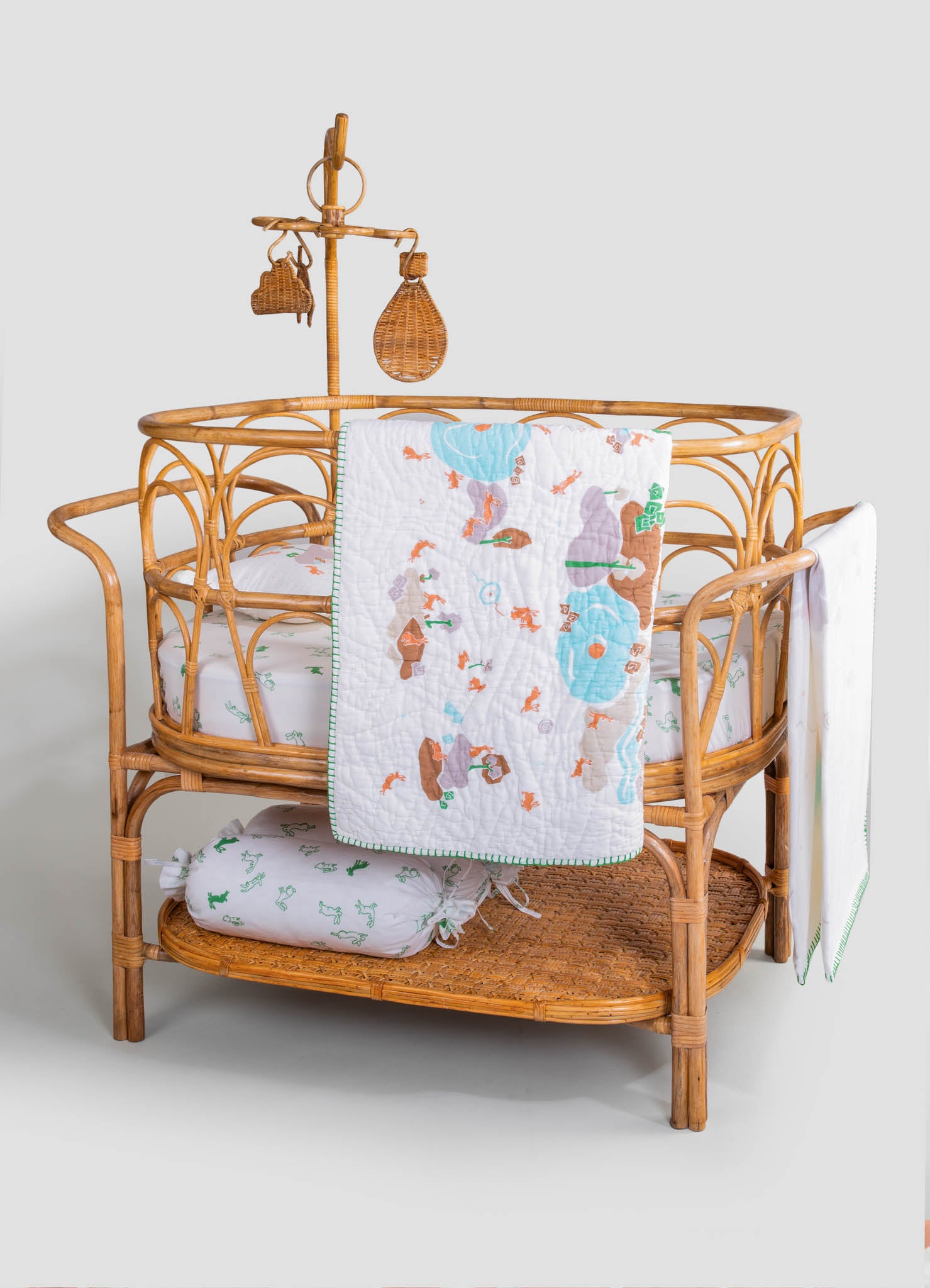 Wicker bassinet with an oval shape, featuring intricate woven rattan sides and a matching woven lower shelf. It is decorated with embroidered floral and animal motifs in soft pastel colors on a white fabric lining, accompanied by a quilt and a pillow with green embroidered designs.