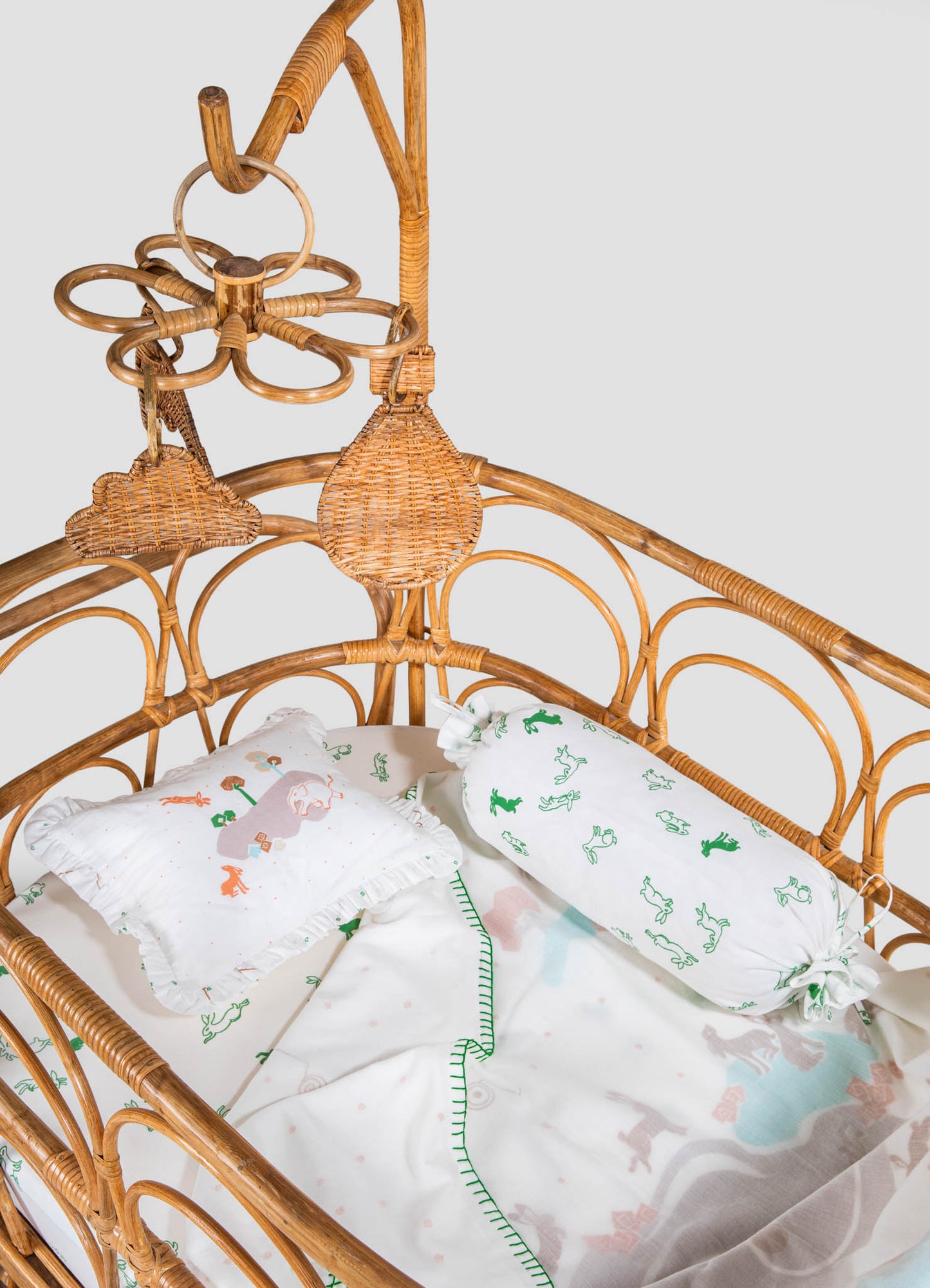 A rattan crib with a curved design, featuring woven natural wicker elements and decorative elements like a flower-shaped hanging ornament. The bedding includes a white sheet with colorful animal and dinosaur motifs, and a pillow with a ruffled border.