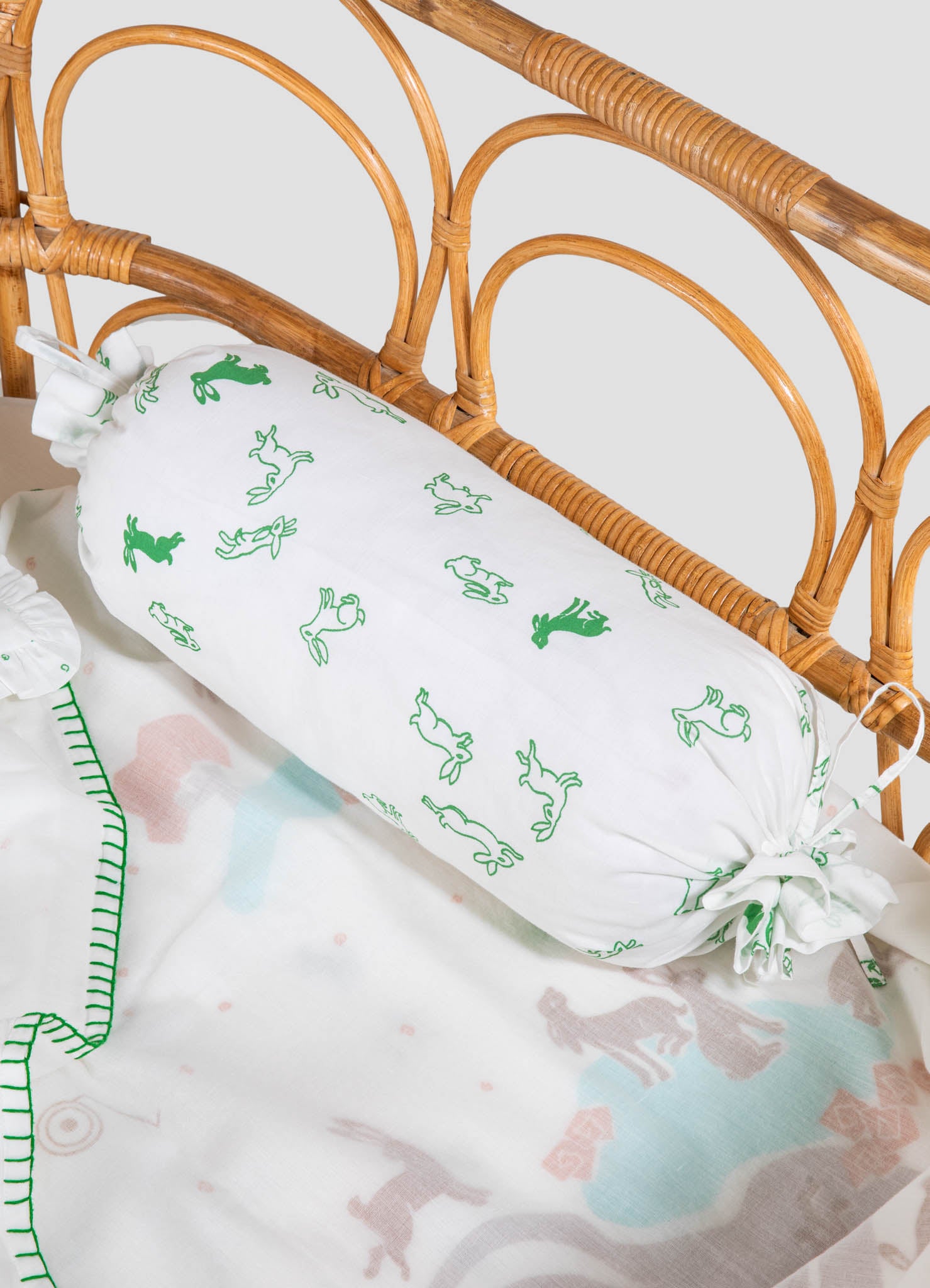 Cotton baby pillow with green animal print motifs placed on a wicker crib bed with a curved rattan frame. The bedding features playful multicolored dinosaur illustrations.