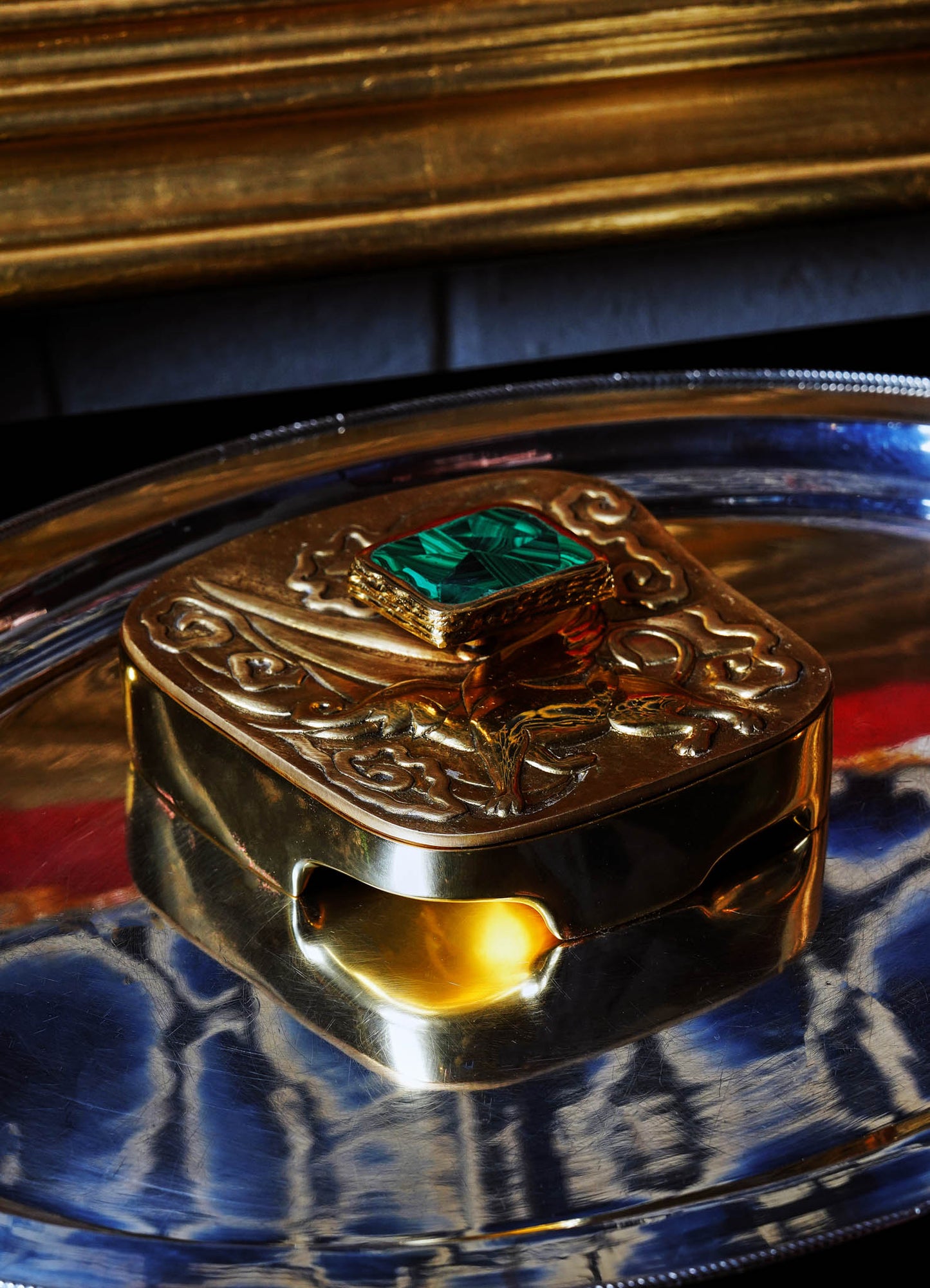 A decorative brass box with ornate embossed patterns and a square green gemstone on the lid, placed on a round metallic tray with reflective surfaces. The box has a vintage style with a polished finish.