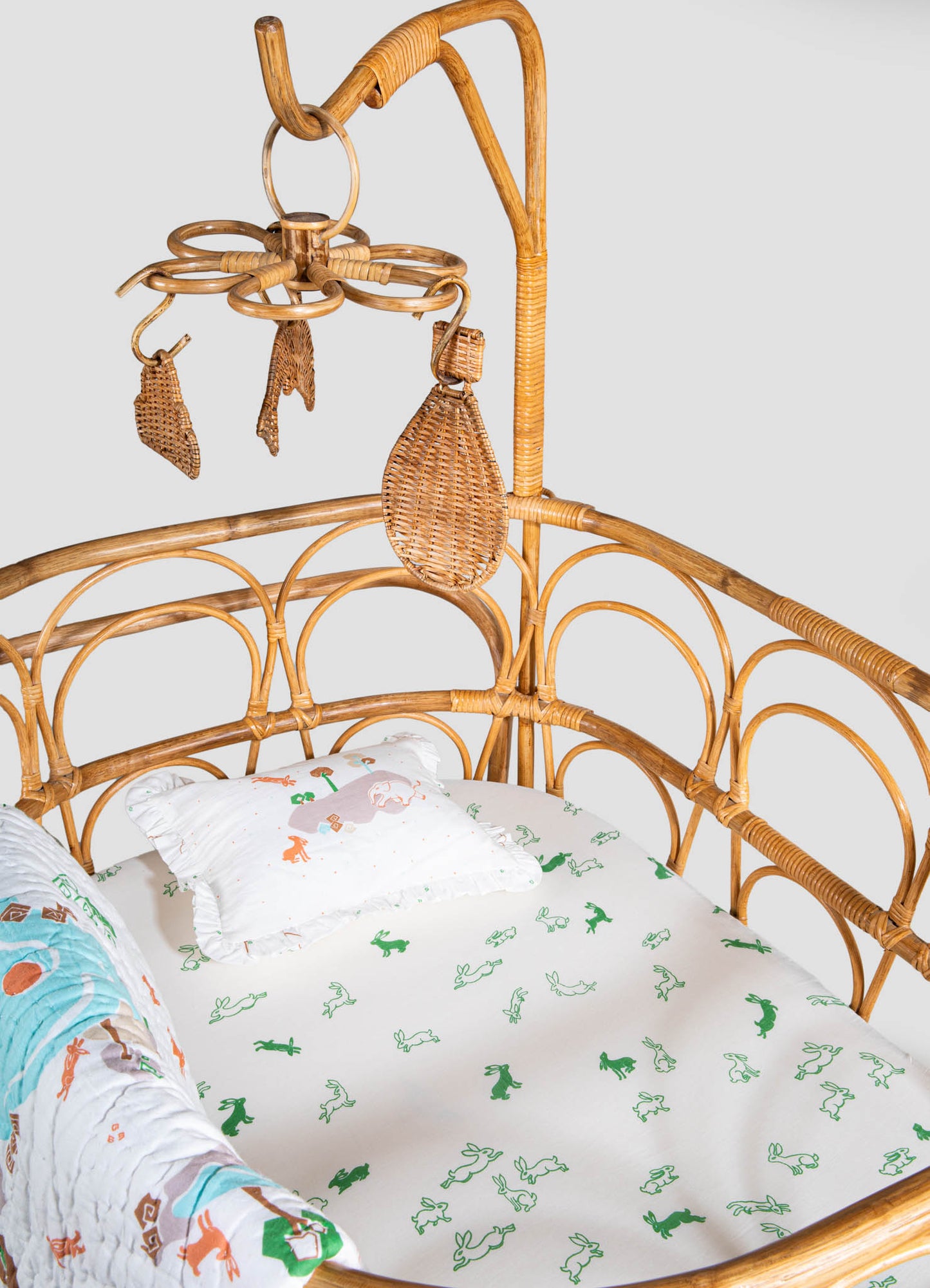 Bamboo crib with a curved rattan headboard and side rails, featuring a white fitted sheet with green animal motifs and matching ruffled pillowcase. A small quilt with colorful jungle animal prints is draped over the side of the crib.