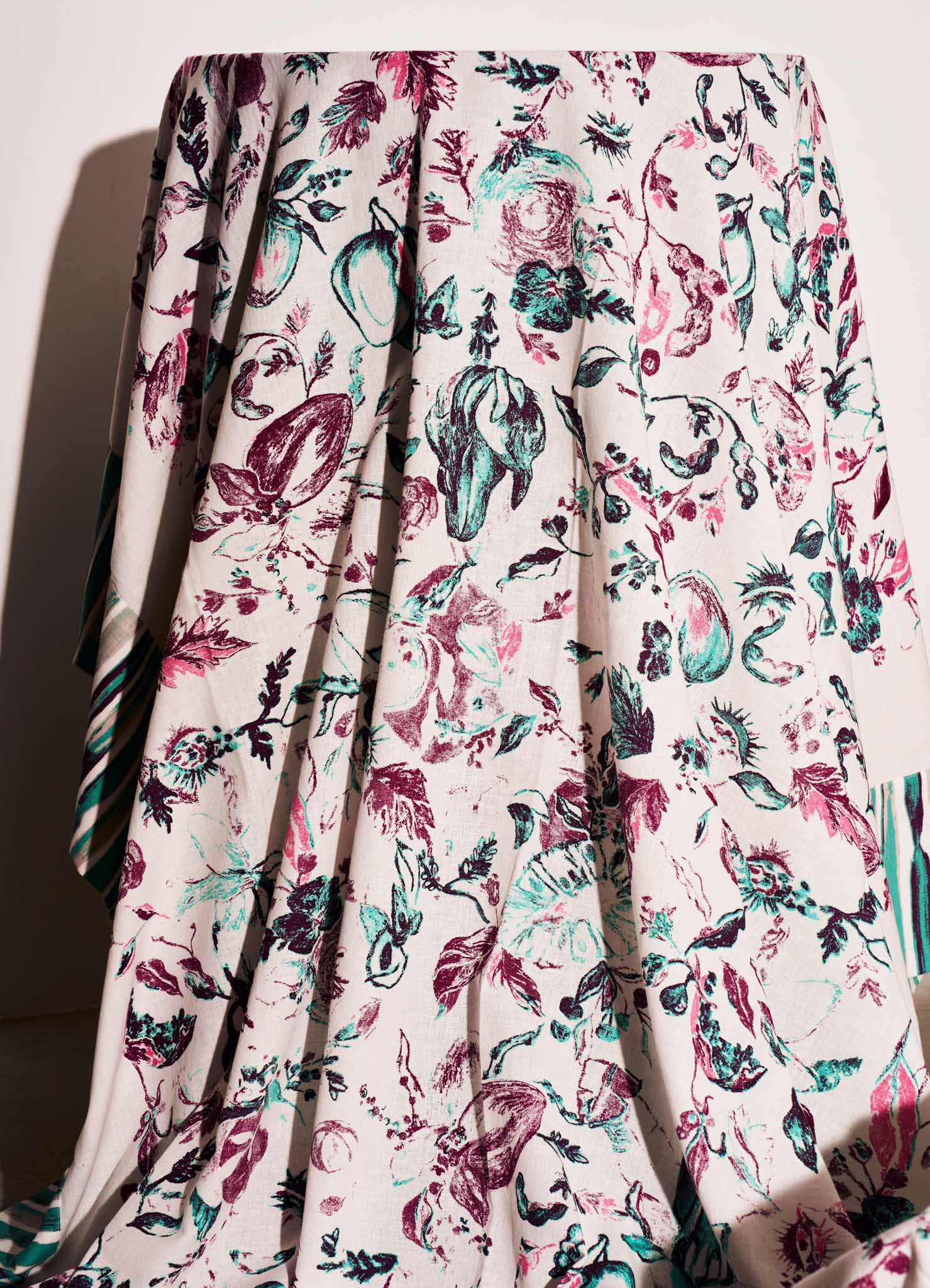 A floral-patterned fabric with a light background, featuring illustrations of various fruits, leaves, and botanical elements in shades of pink, purple, teal, and black. The material appears to be lightweight and has a pleated, flowing drape.