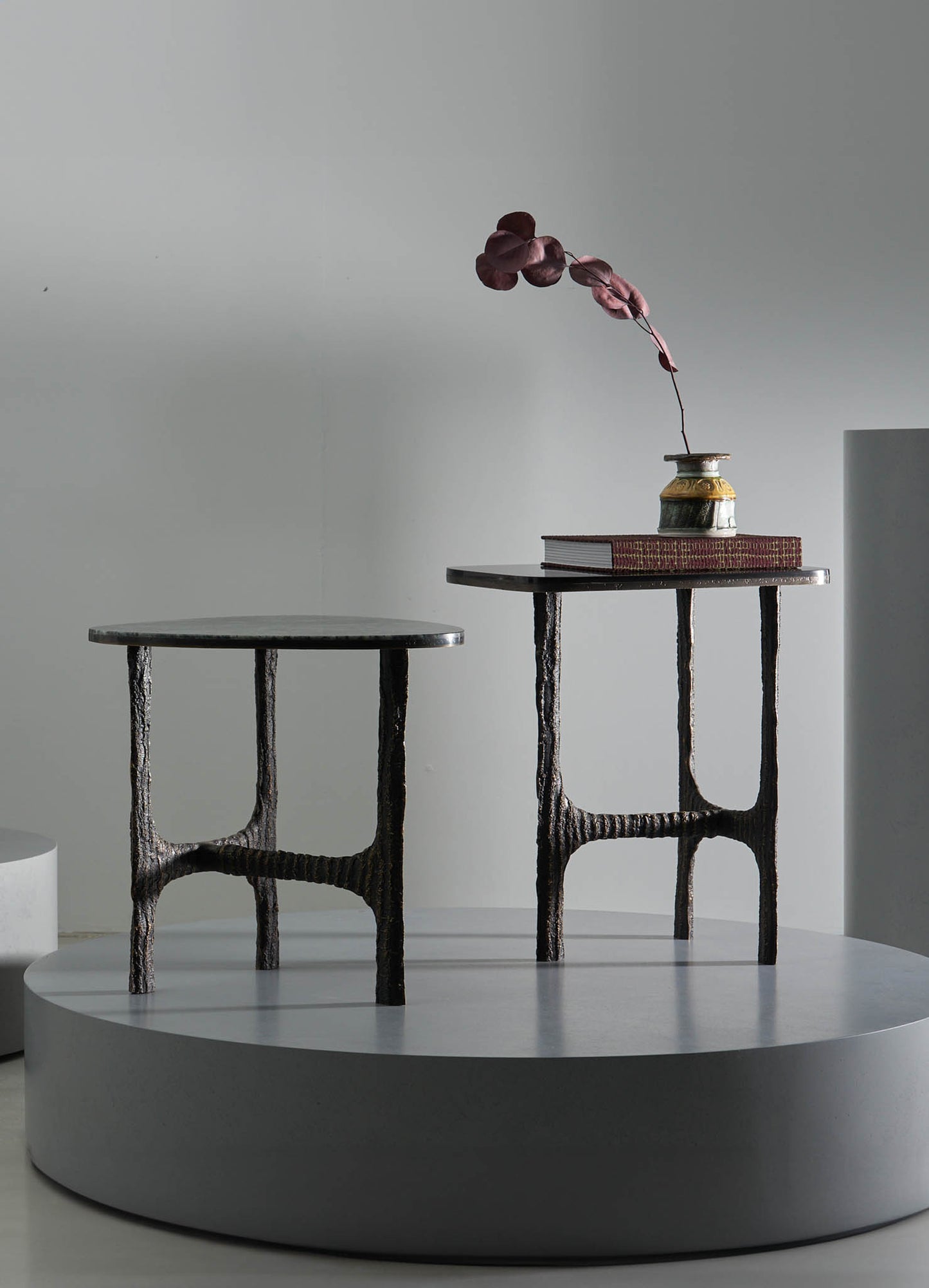 Two small tables with textured, dark metal frames and smooth, flat tabletops. One table has a rounded top, while the other has a rectangular top with slightly beveled edges, both set on a minimalist white platform. The rectangular table features a ceramic vase with a gold accent and a closed book, with a single branch of deep red leaves extending from the vase.