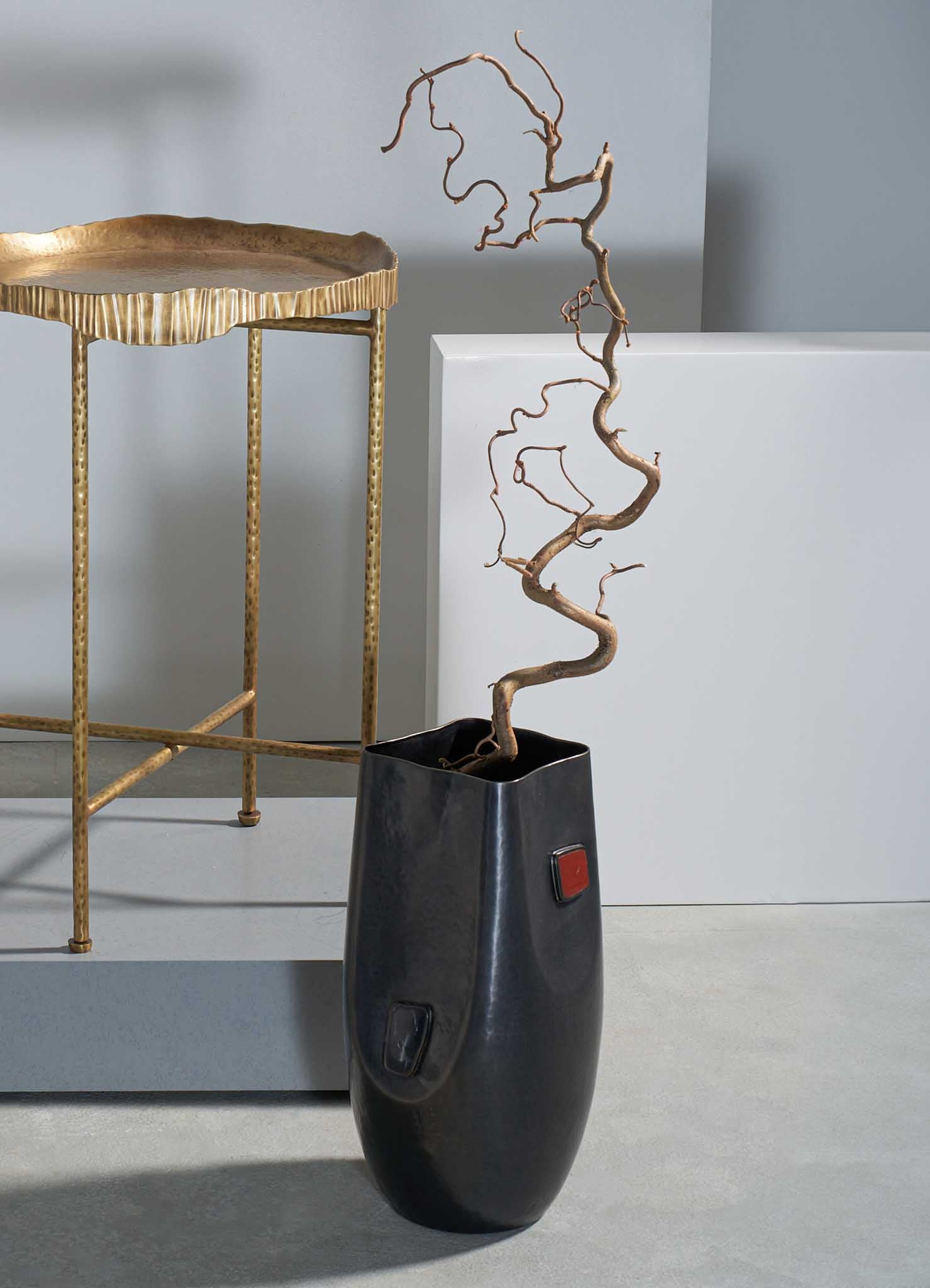 A tall, black ceramic vase with a smooth finish holds a twisted, leafless branch with a natural, organic shape. The scene also includes a gold metal side table with a scalloped edge and textured surface, set against a light gray wall.