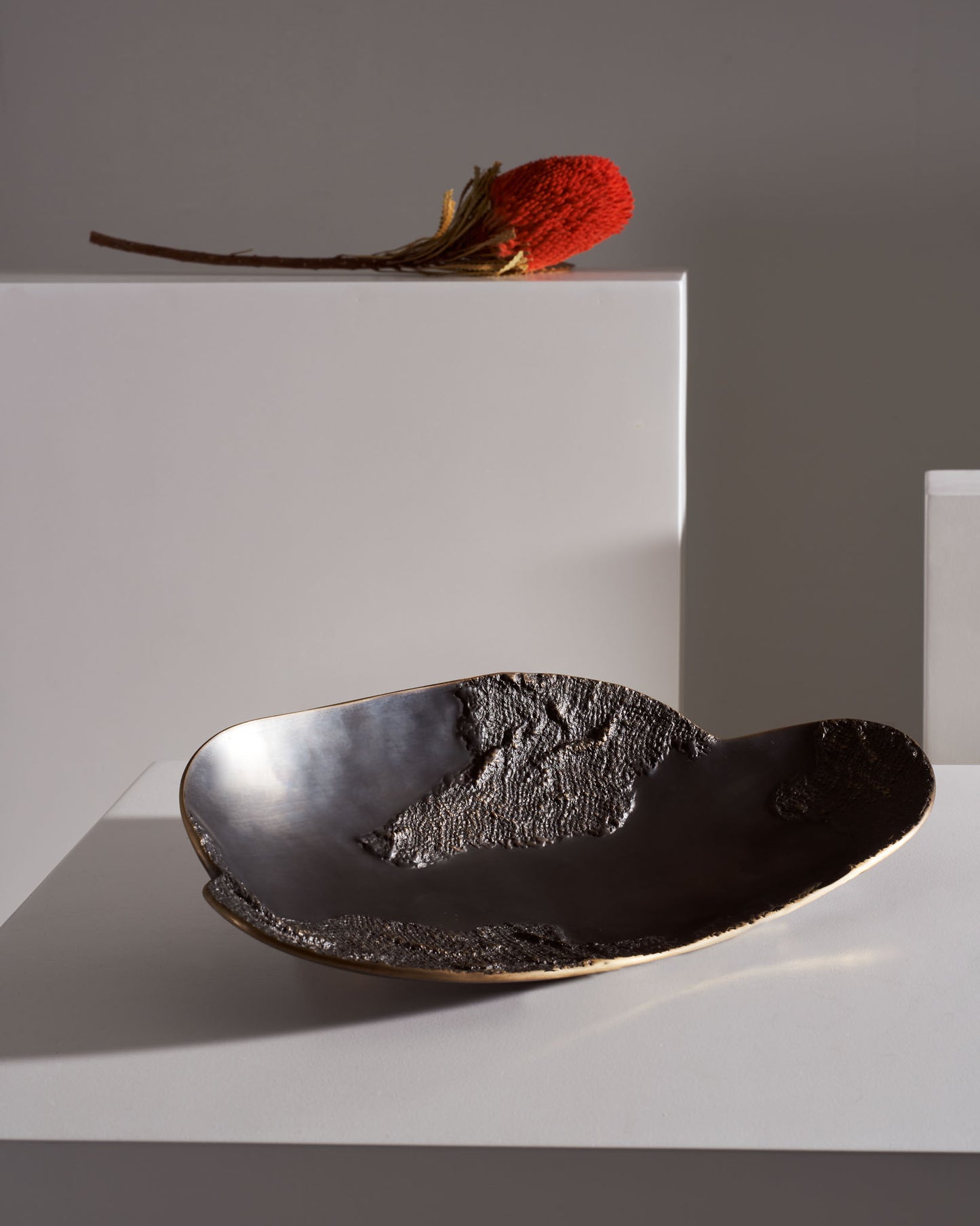 A black ceramic dish with an irregular, textured surface and a smooth, glossy interior, placed on a white tabletop. A red dried flower with a brown stem rests on a white rectangular block in the background, with a neutral gray wall behind.