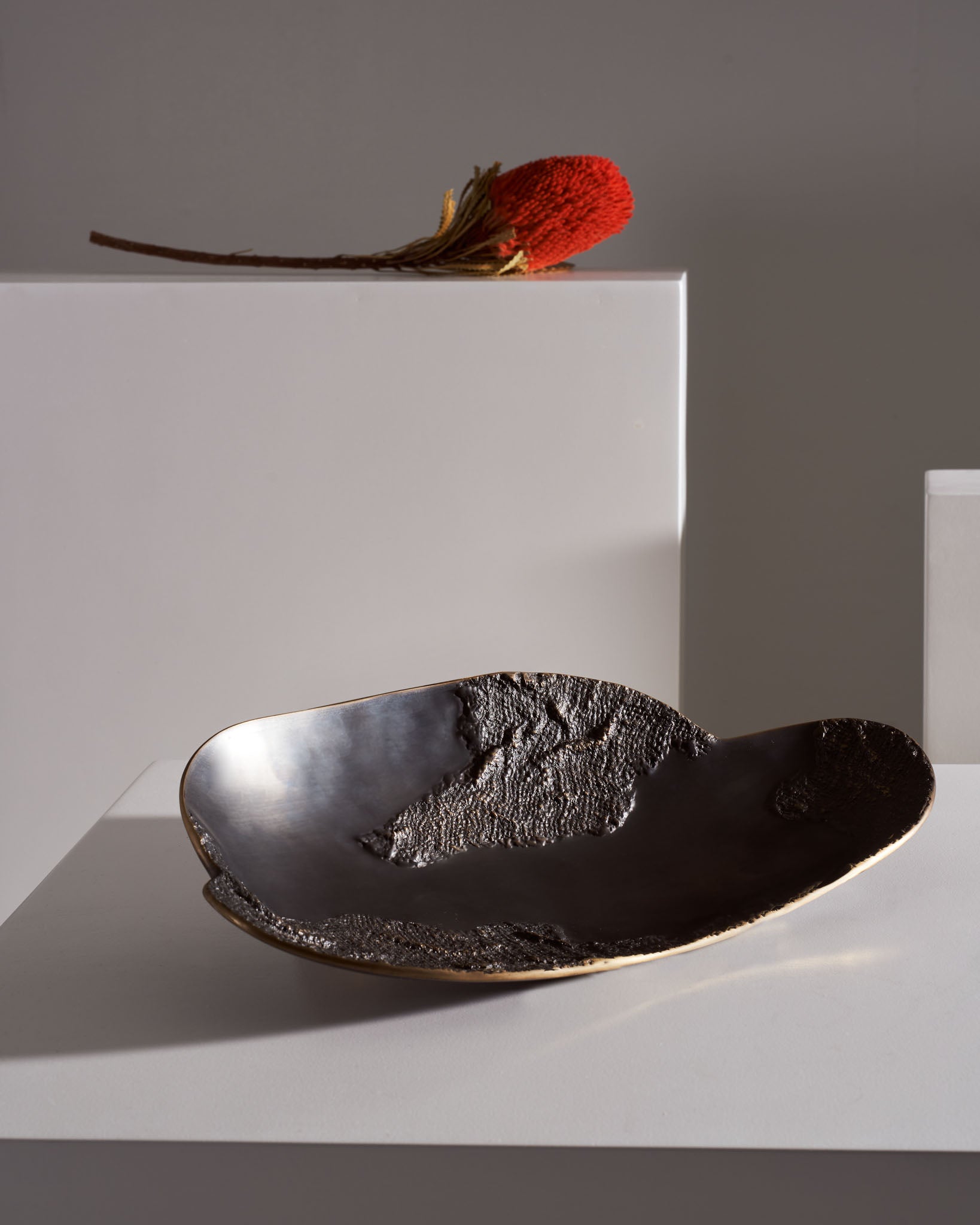 A black ceramic dish with an irregular, textured surface and a smooth, glossy interior, placed on a white tabletop. A red dried flower with a brown stem rests on a white rectangular block in the background, with a neutral gray wall behind.