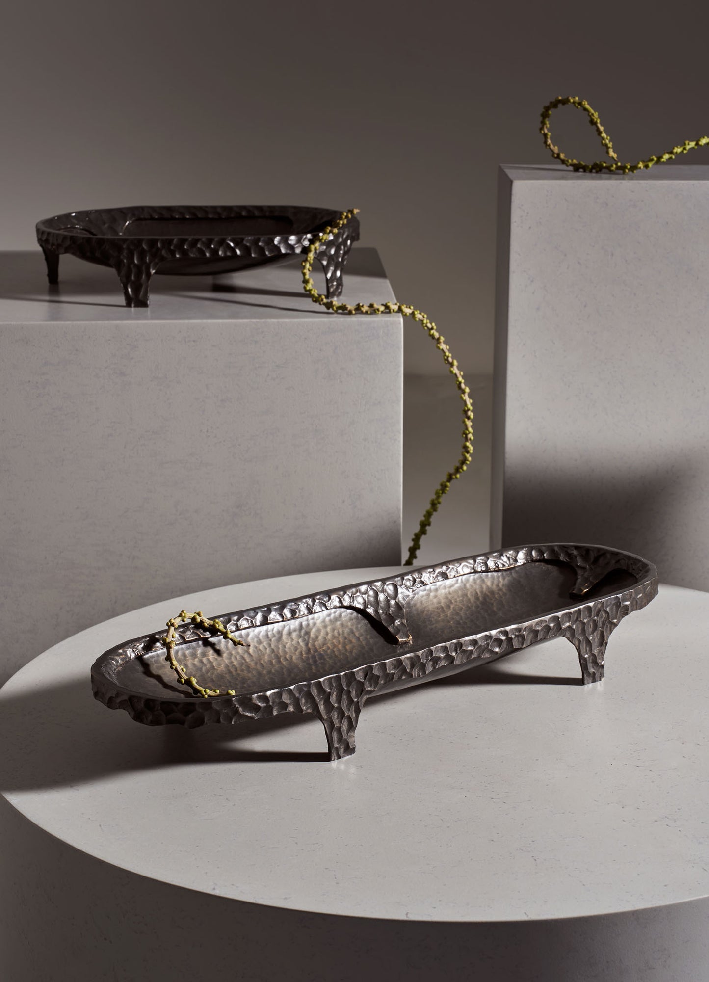 Hammered metal trays with a textured, dark finish and elongated, oval shape, displayed on white pedestals. The trays are complemented by decorative beaded strings in yellow-green tones.
