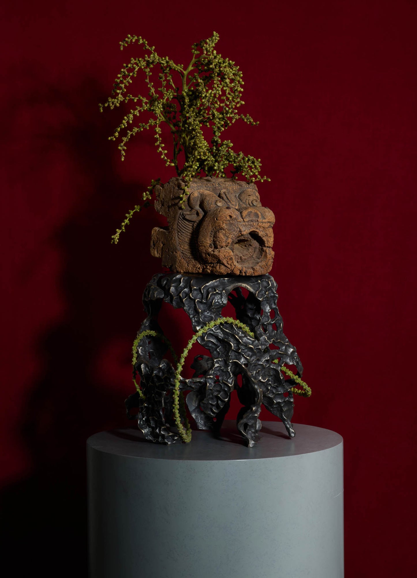 Decorative sculpture featuring an intricately carved wooden face with a fierce expression, mounted on a black, textured, irregularly shaped base with green plant elements arranged above against a deep red background.