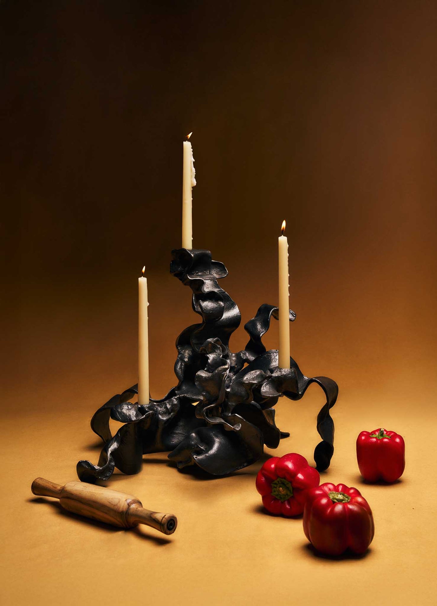 A sculptural candle holder with an abstract, twisted black metal design, holding three tall, lit beige candles. The setup is complemented by three red bell peppers and a wooden rolling pin placed on a warm-toned surface, against a gradient brown background.