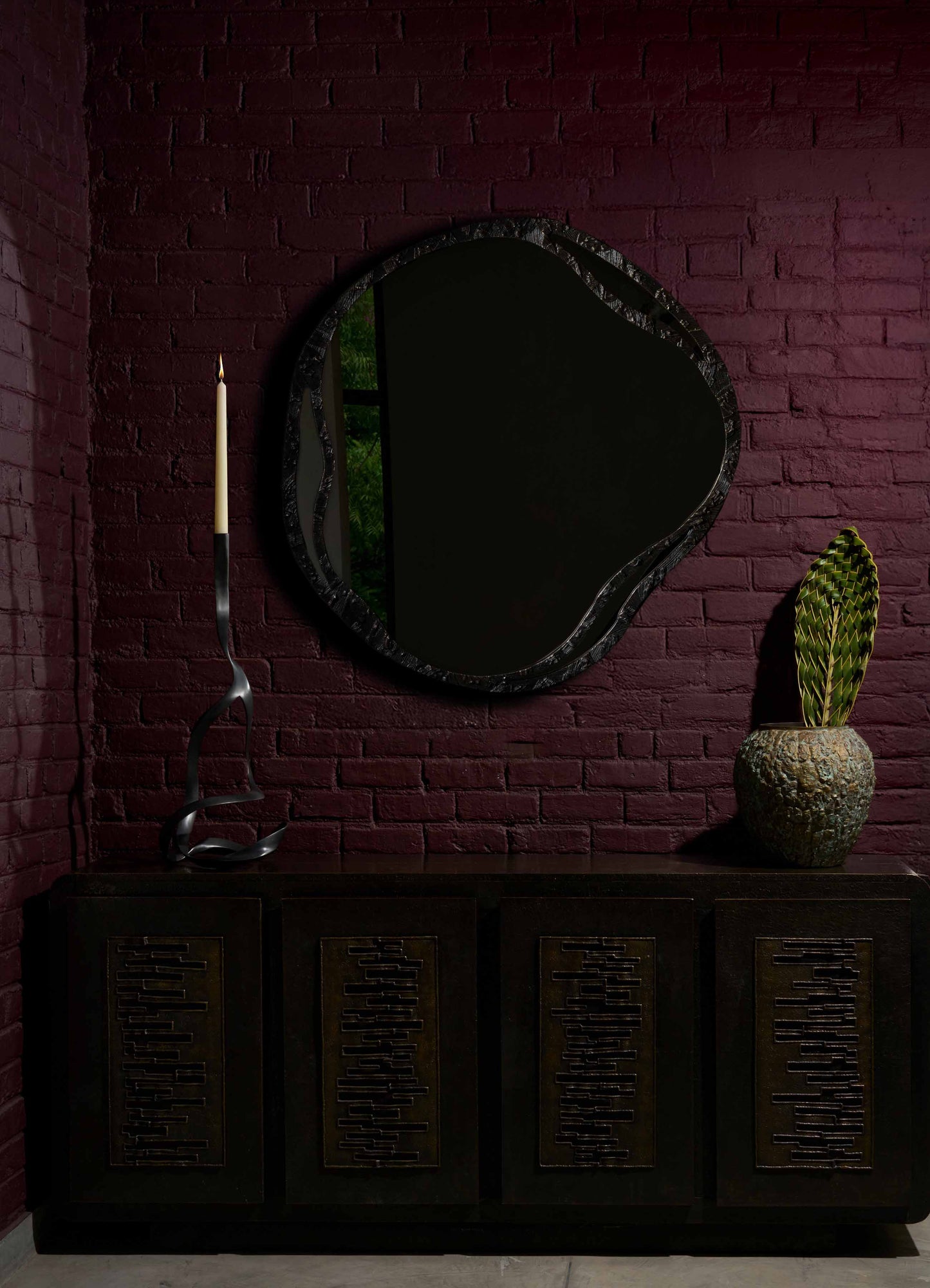 A black wooden sideboard with geometric cutout patterns in a dark interior setting, topped with a tall, sculptural black candle holder with a curling design, a large textured ceramic vase with green variegated leaves, and a large irregularly shaped mirror with a black frame on a deep red brick wall.
