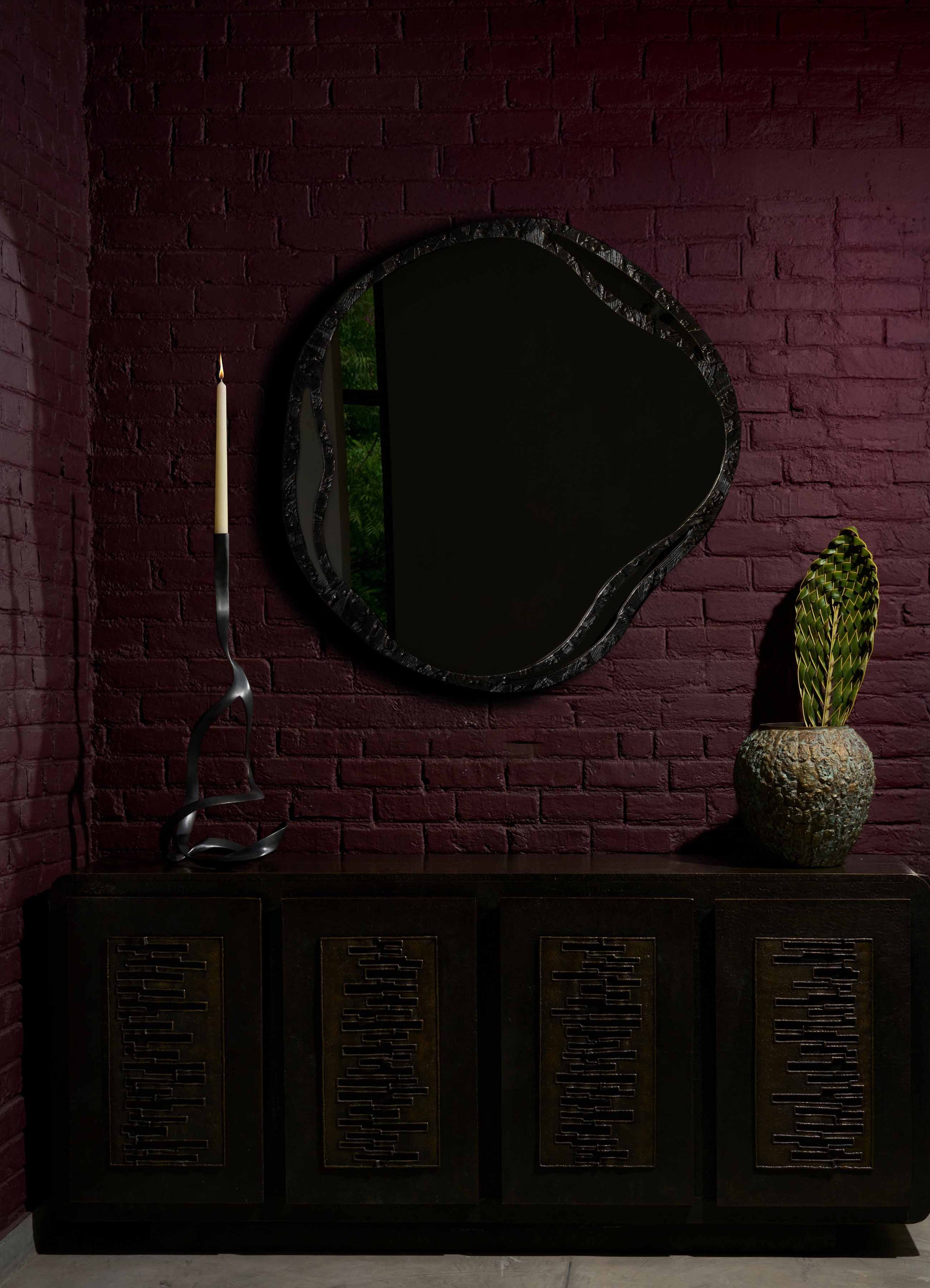 A black wooden sideboard with geometric cutout patterns in a dark interior setting, topped with a tall, sculptural black candle holder with a curling design, a large textured ceramic vase with green variegated leaves, and a large irregularly shaped mirror with a black frame on a deep red brick wall.