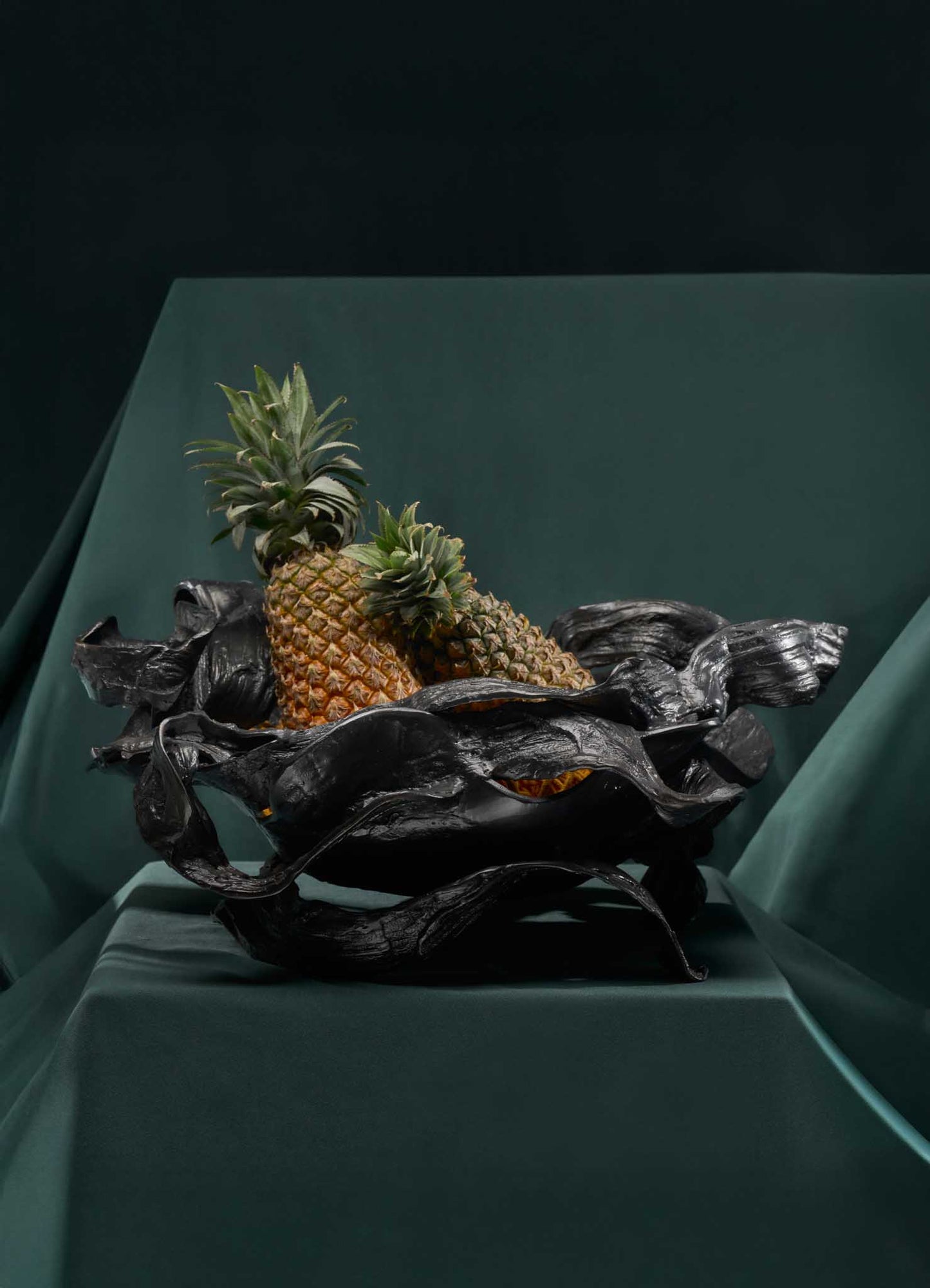 A black sculptural bowl with an organic, flowing shape holding three pineapples with green spiky leaves, set on a dark green surface against a dark background.