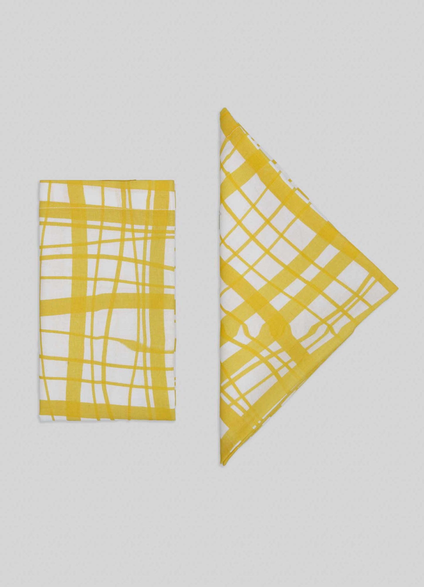 Viya’s Madras Check Yellow Napkins – Set of 4 are displayed on a light gray background, with two white napkins featuring yellow Madras checks folded into rectangle and triangle shapes, highlighting contemporary patterns from the Indian subcontinent.