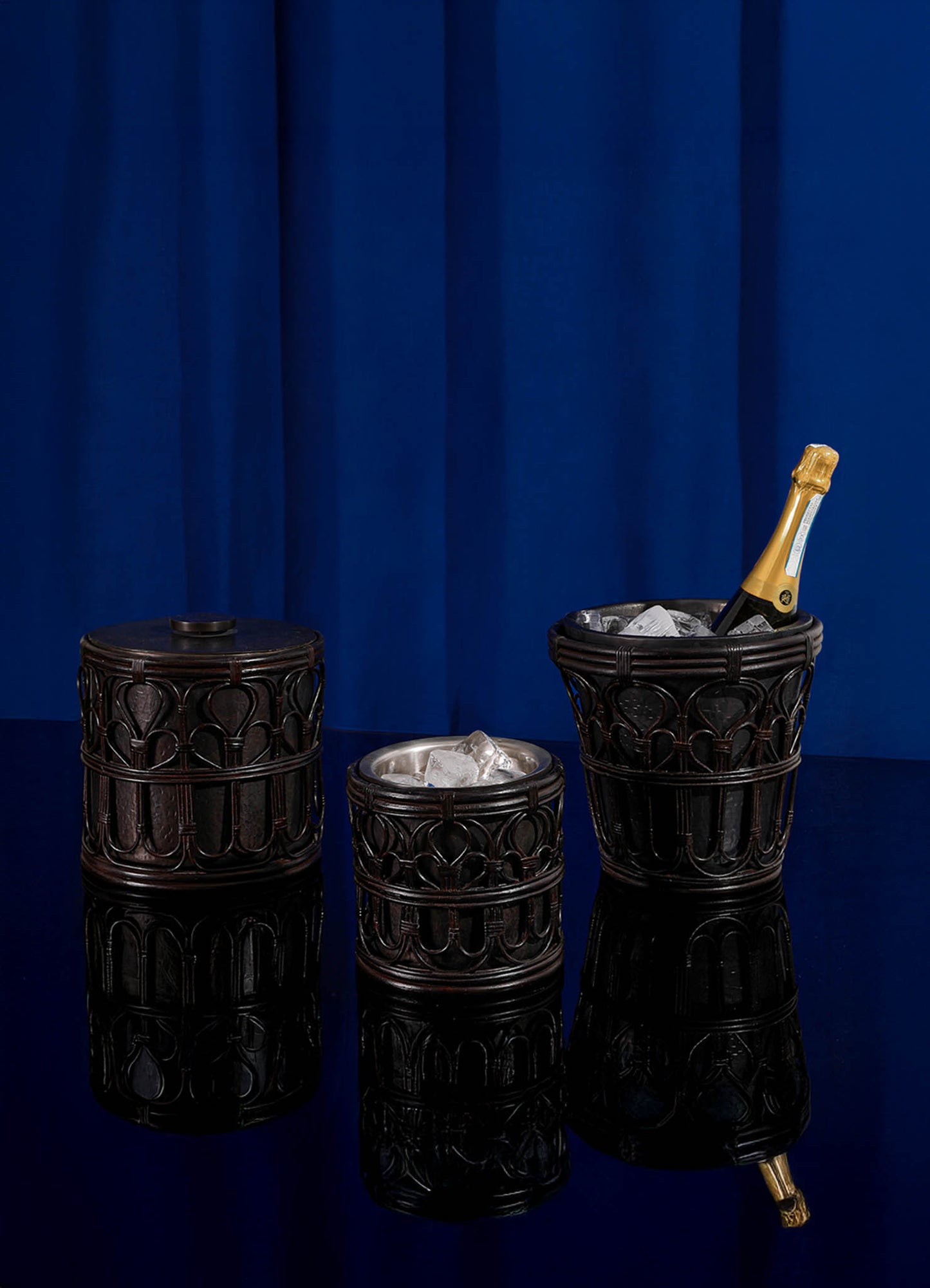 Three dark, ornate VIYA Manipur Ice Bucket Large barware accessories feature cane lattice patterns and hand-beaten brass finishes. Displayed against a blue curtain, one is lidded, one holds ice, and one chills champagne on a reflective surface.