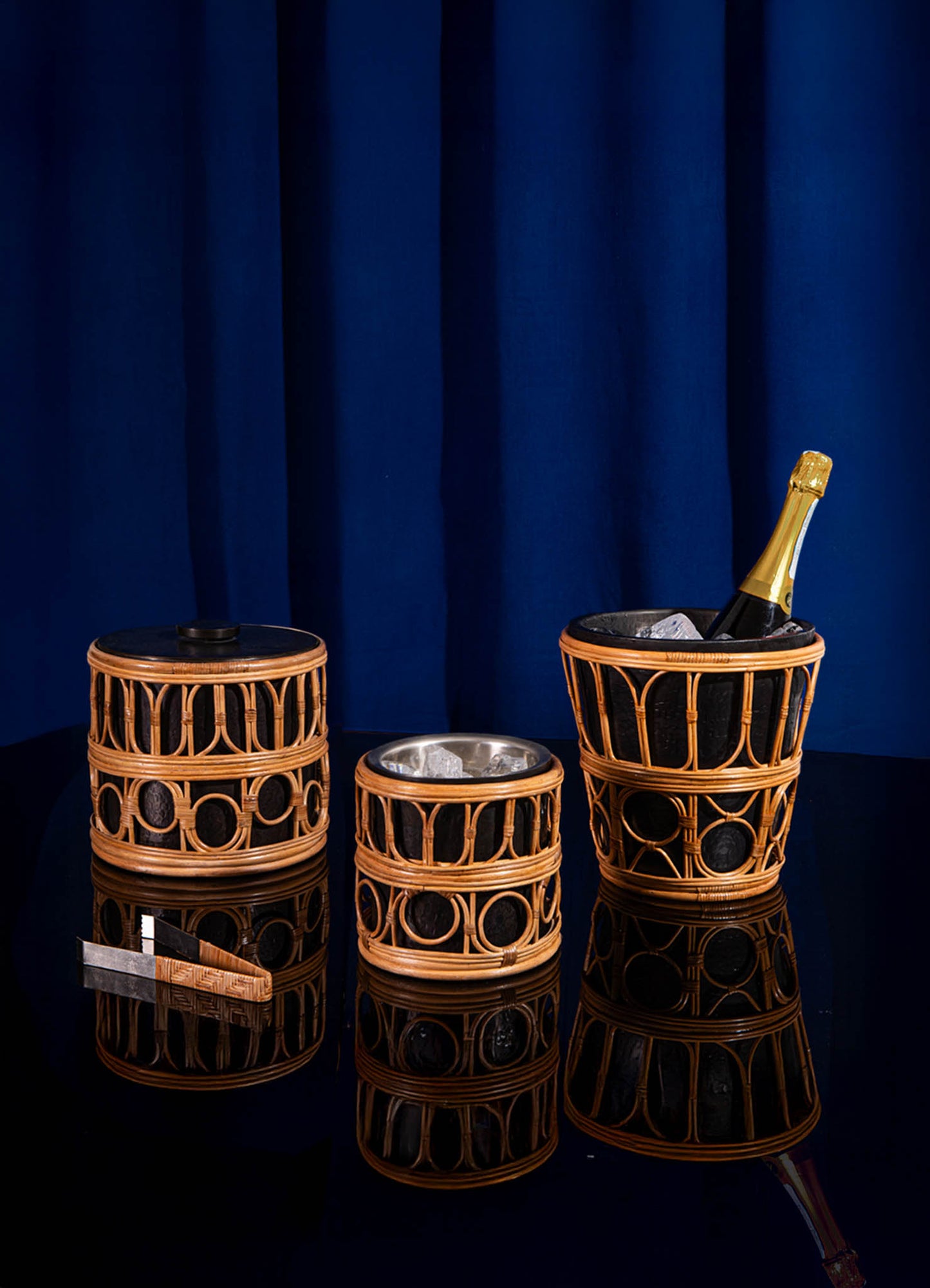 Set of three bamboo and rattan ice buckets with circular cutout motifs and black lining, one containing ice and a champagne bottle, displayed on a glossy black surface against a dark blue fabric background.
