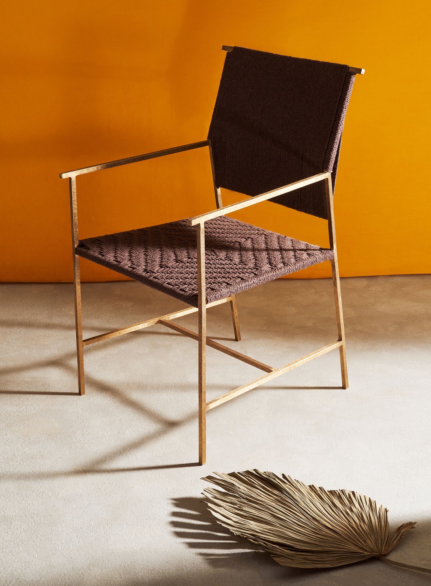 A modern chair with a brass frame, upholstered with dark fabric on the backrest and textured, patterned fabric on the seat. The setting features an orange backdrop, a dried palm leaf on the floor, and a neutral-toned carpet.
