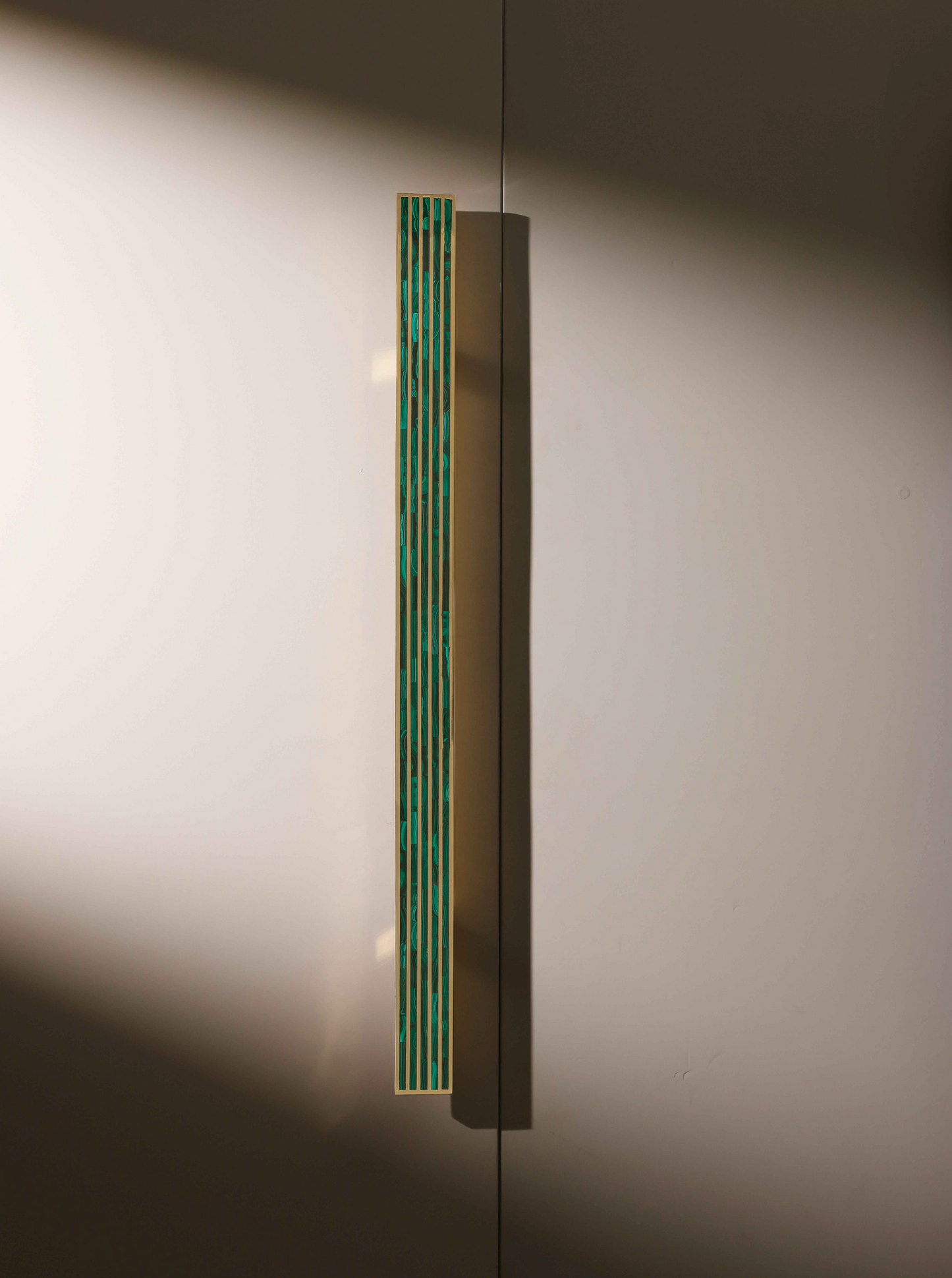 A close-up of a modern cabinet or door handle with a slim, vertical design featuring geometric cutouts and a teal color accents, mounted on a smooth, beige surface.