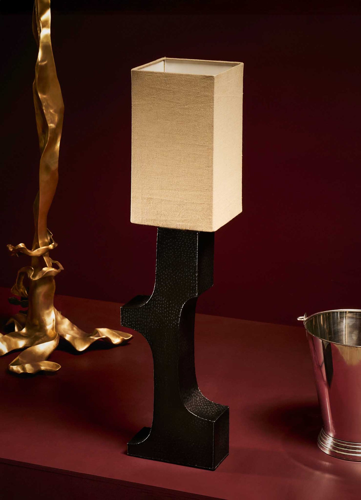 A table lamp with a rectangular beige fabric lampshade and a black textured sculptural base, displayed on a burgundy surface.
