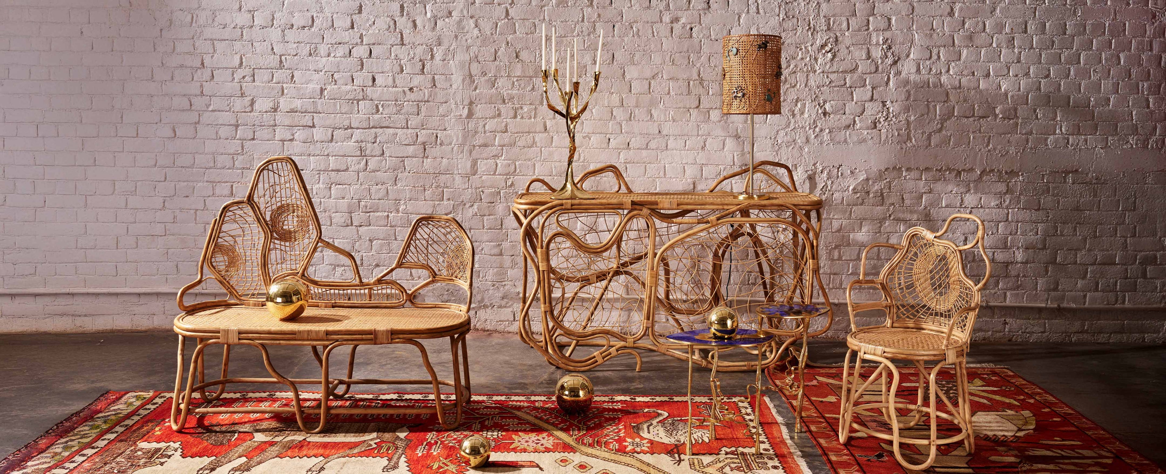 Rattan furniture set including armchairs and a side table with intricate woven patterns, complemented by metallic gold spheres and a small glass table with blue accents. The pieces feature natural wood tones, crafted in a handcrafted, eclectic style, placed on a red patterned rug against a white brick wall.