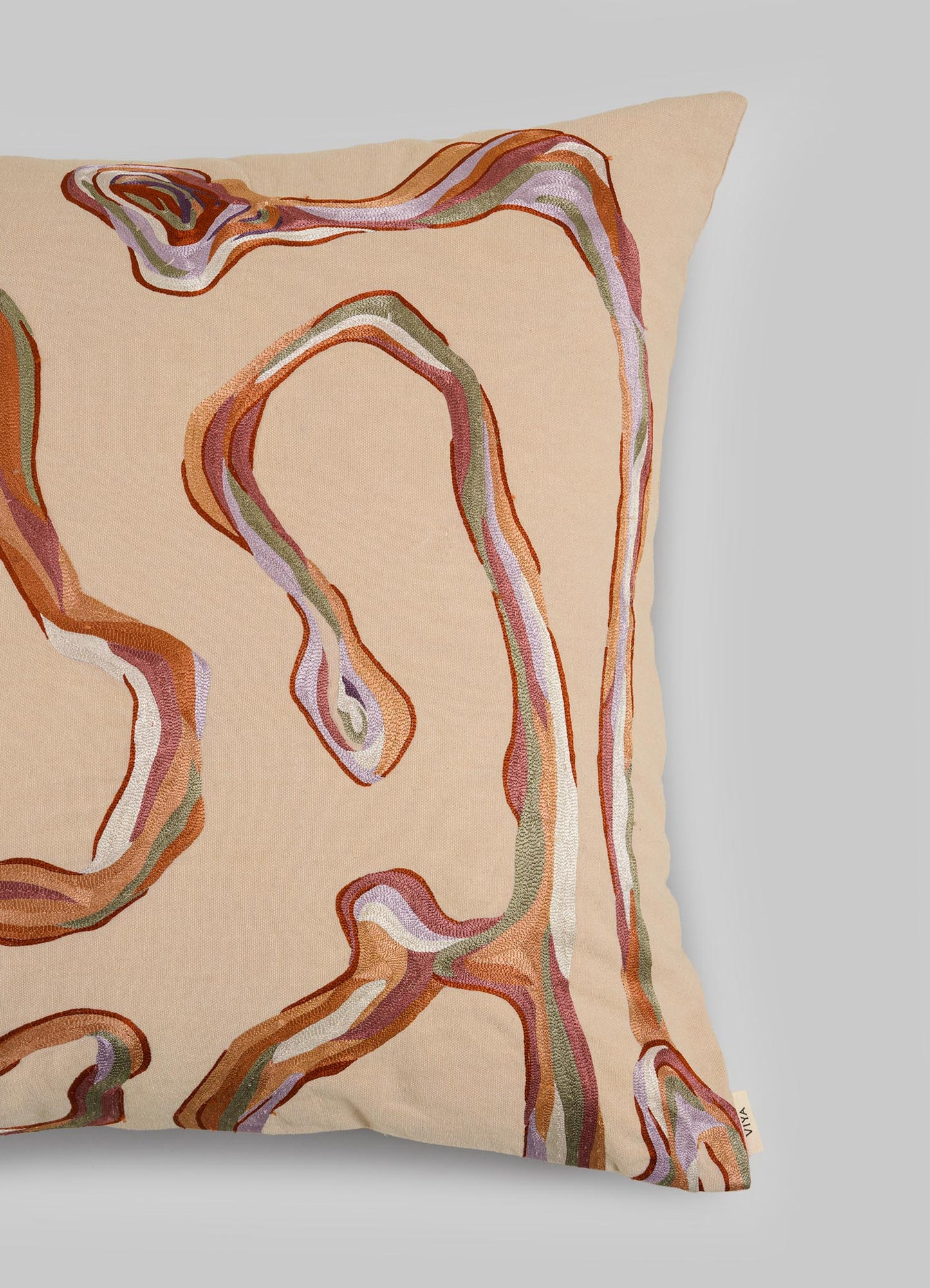 The Viya Tutti Frutti Salmon Pink Cushion Cover (24"x24") features abstract, wavy hand-embroidered lines in brown, rust, pink, green, and white on a beige background—its fluid pattern inspired by Cartier’s Tutti Frutti.