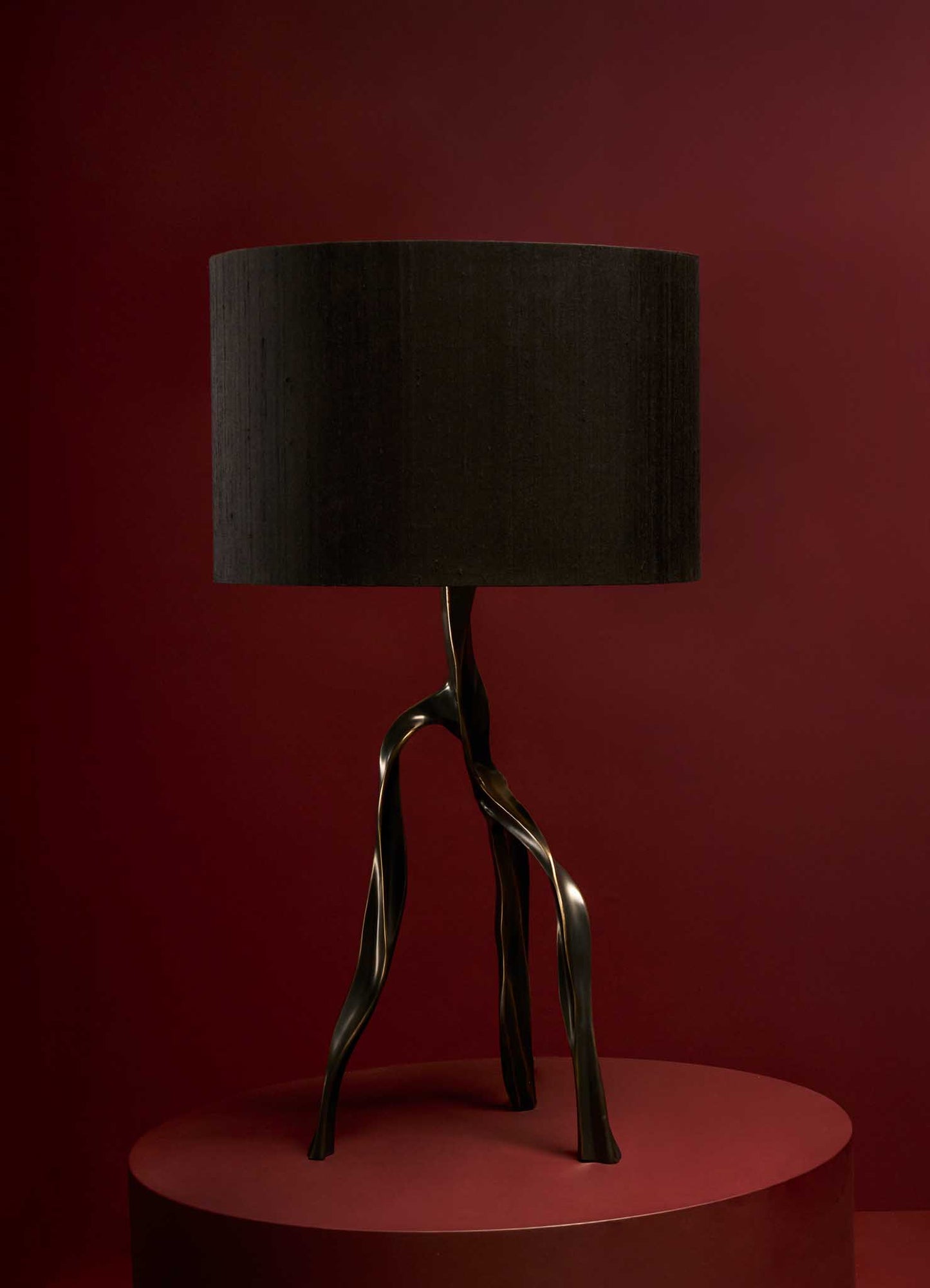 Decorative table lamp with a sculptural metal base resembling twisted branches and a cylindrical black fabric shade, set against a dark red background.
