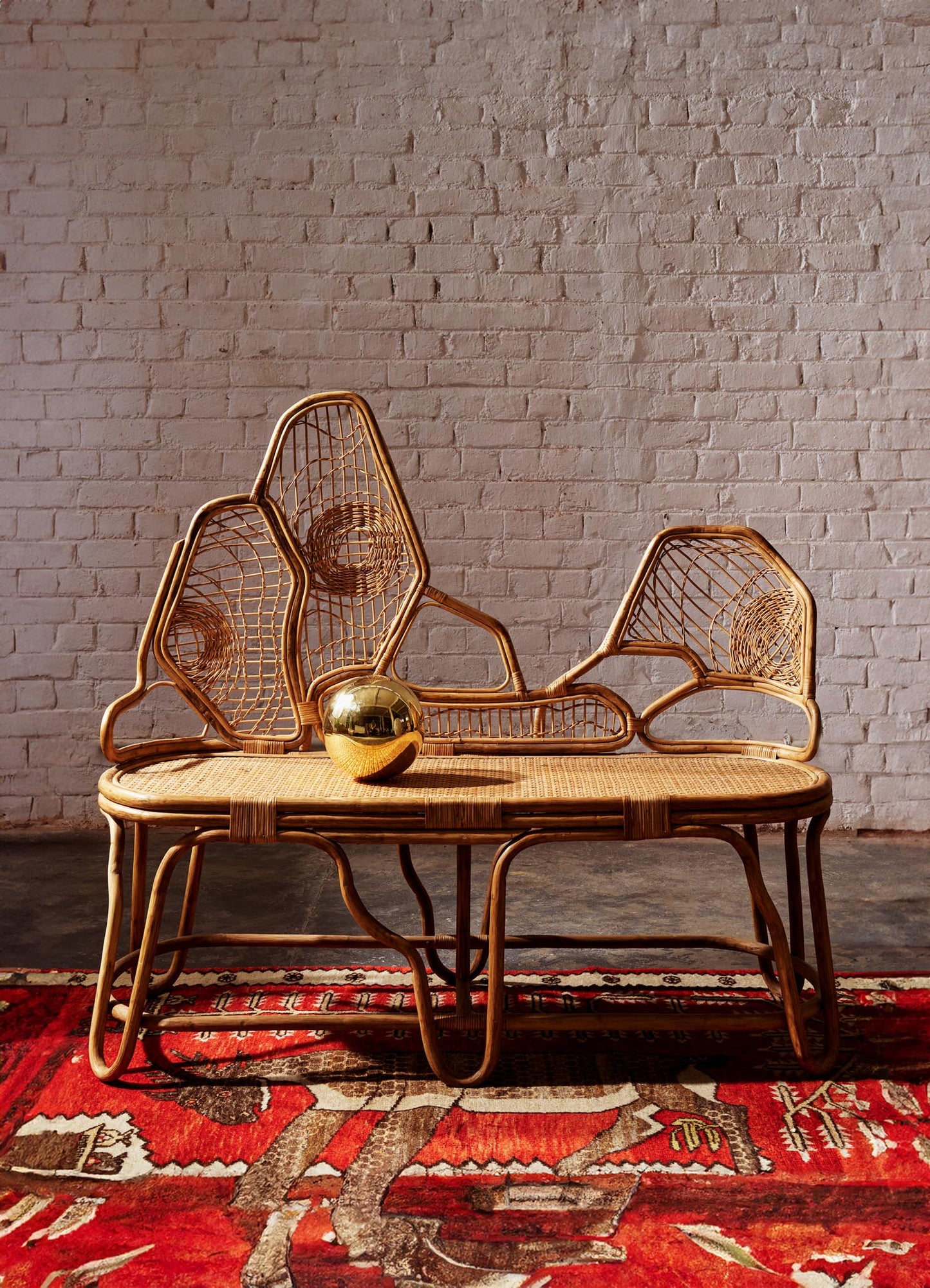 Rattan bench with a woven seat and decorative backrest featuring geometric and spiral motifs, accompanied by a reflective gold spherical object. The bench is set against a textured pink brick wall on a red patterned rug.
