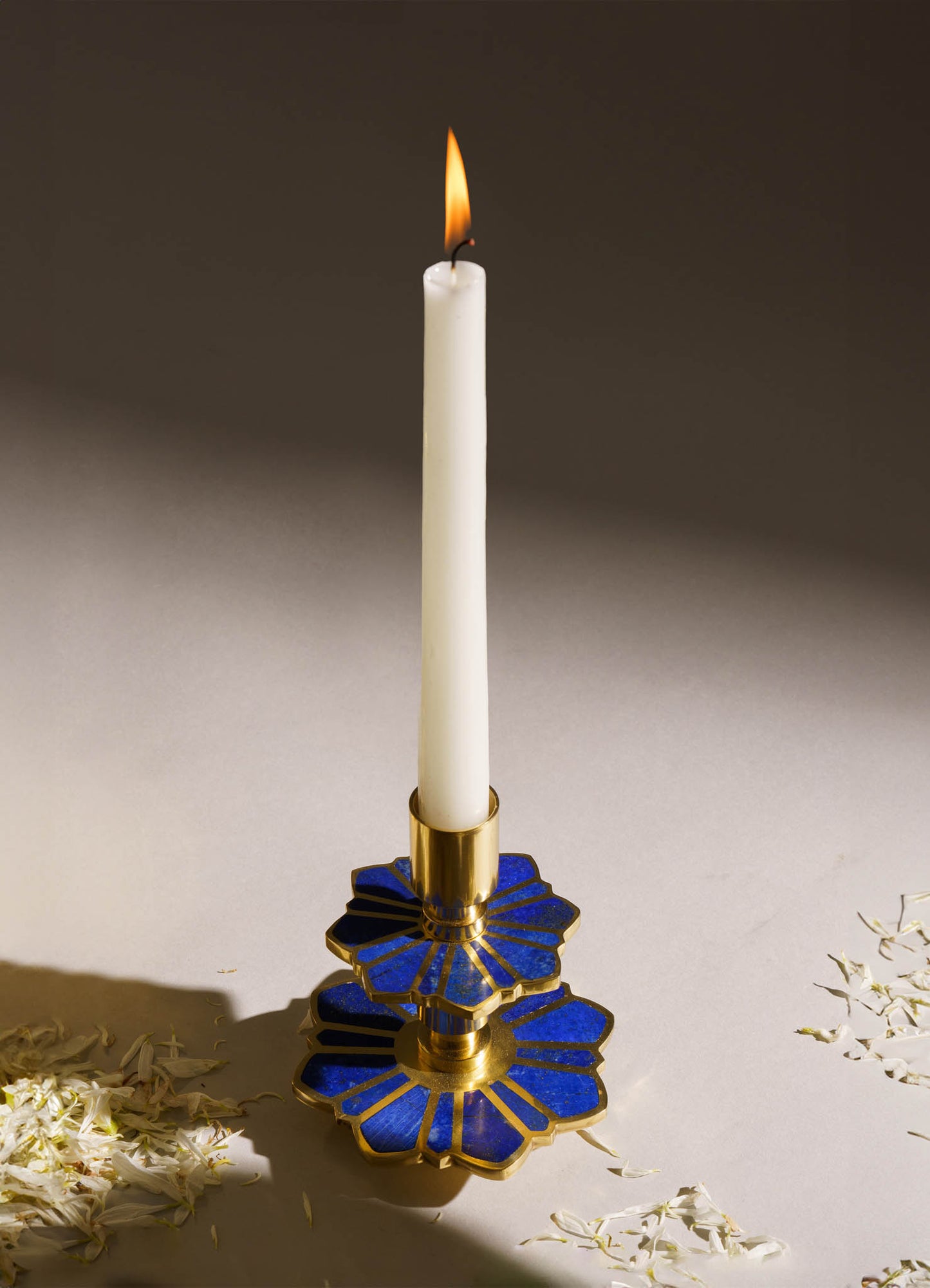 A tall white candlestick with a lit candle features a base and decorative sections in metallic gold and deep blue, with a geometric floral motif. The candle holder combines metal and ceramic materials, presenting an elegant and contemporary style.