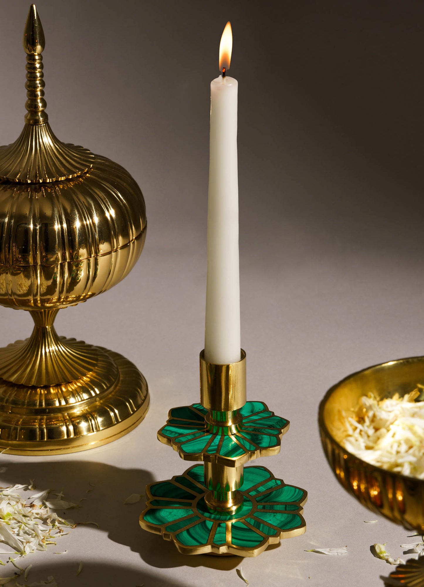 A tall white taper candle with a flickering flame, mounted on a decorative green and gold malachite-style metal candlestick holder. The scene also features a gold ornamental container with a lid and a finial, and a gold bowl filled with shredded material, all set against a neutral background.
