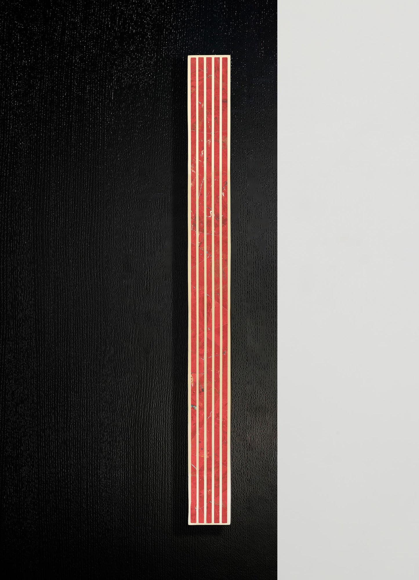 A vertical mosaic tile with red and beige geometric patterns, mounted on a black background with a neutral-colored wall on the right side.