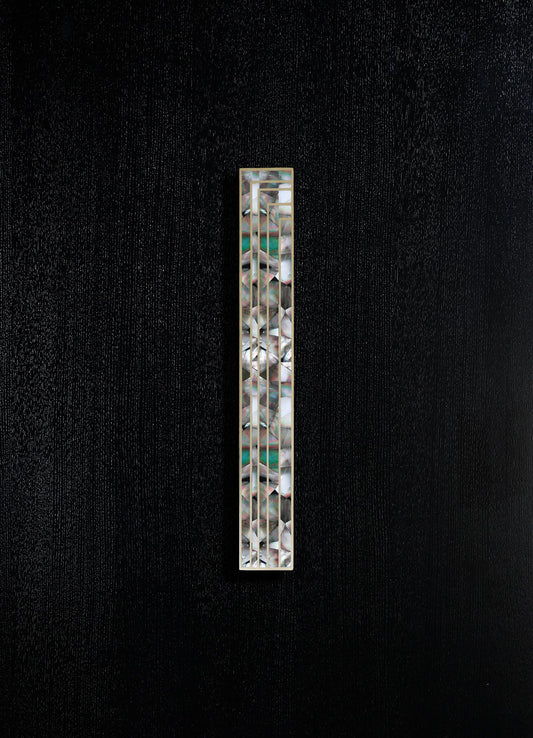 A slim rectangular decorative object featuring a geometric mosaic pattern with iridescent shell-like pieces and metallic accents, set against a textured black background.